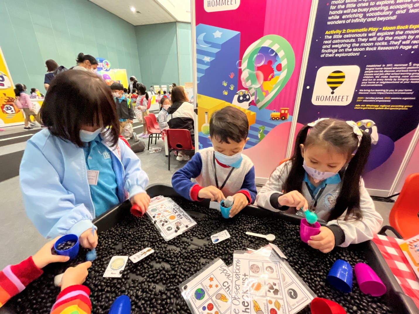 Three young children wearing masks sort black beans in a tray at a Hommeet educational activity event.
