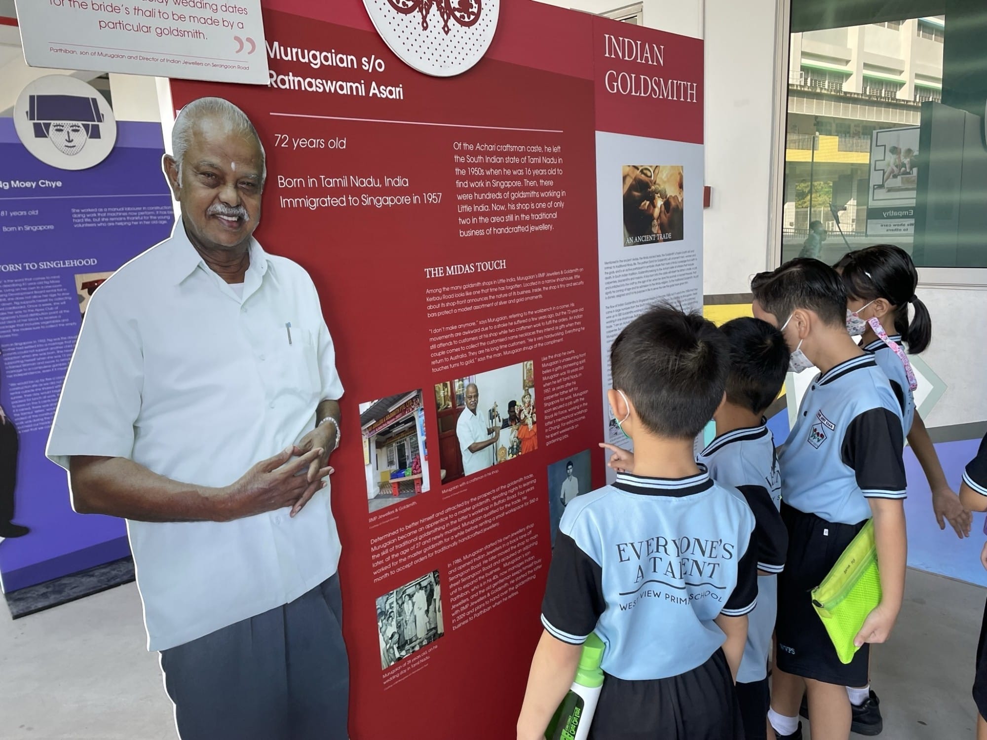 Students in blue uniforms view an outdoor exhibit panel about Indian Goldsmith Murugaian, born in Tamil Nadu.