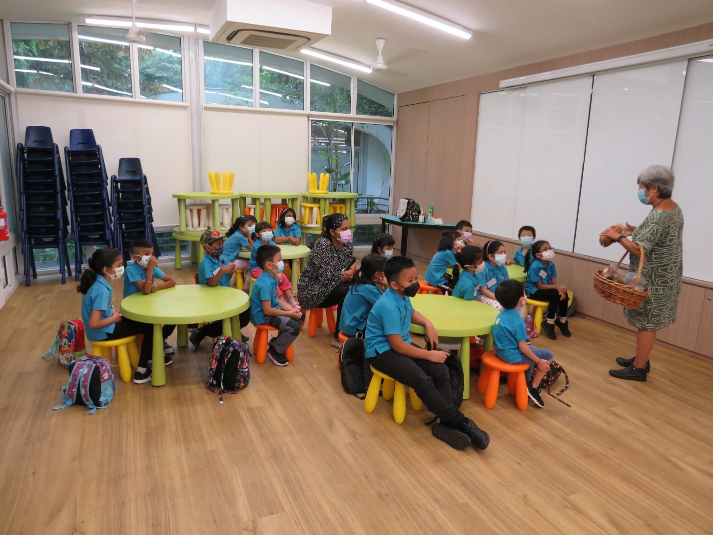 Young children in blue uniforms and masks sit at colorful tables in a classroom, facing an adult holding a basket.