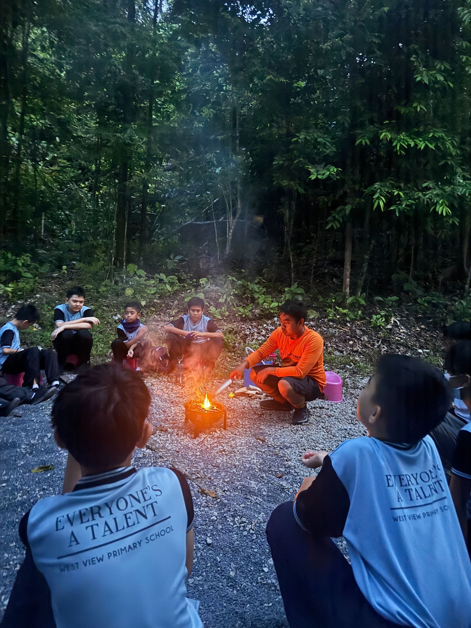 West View Primary School students sit around a small campfire on a gravel path surrounded by dense forest.