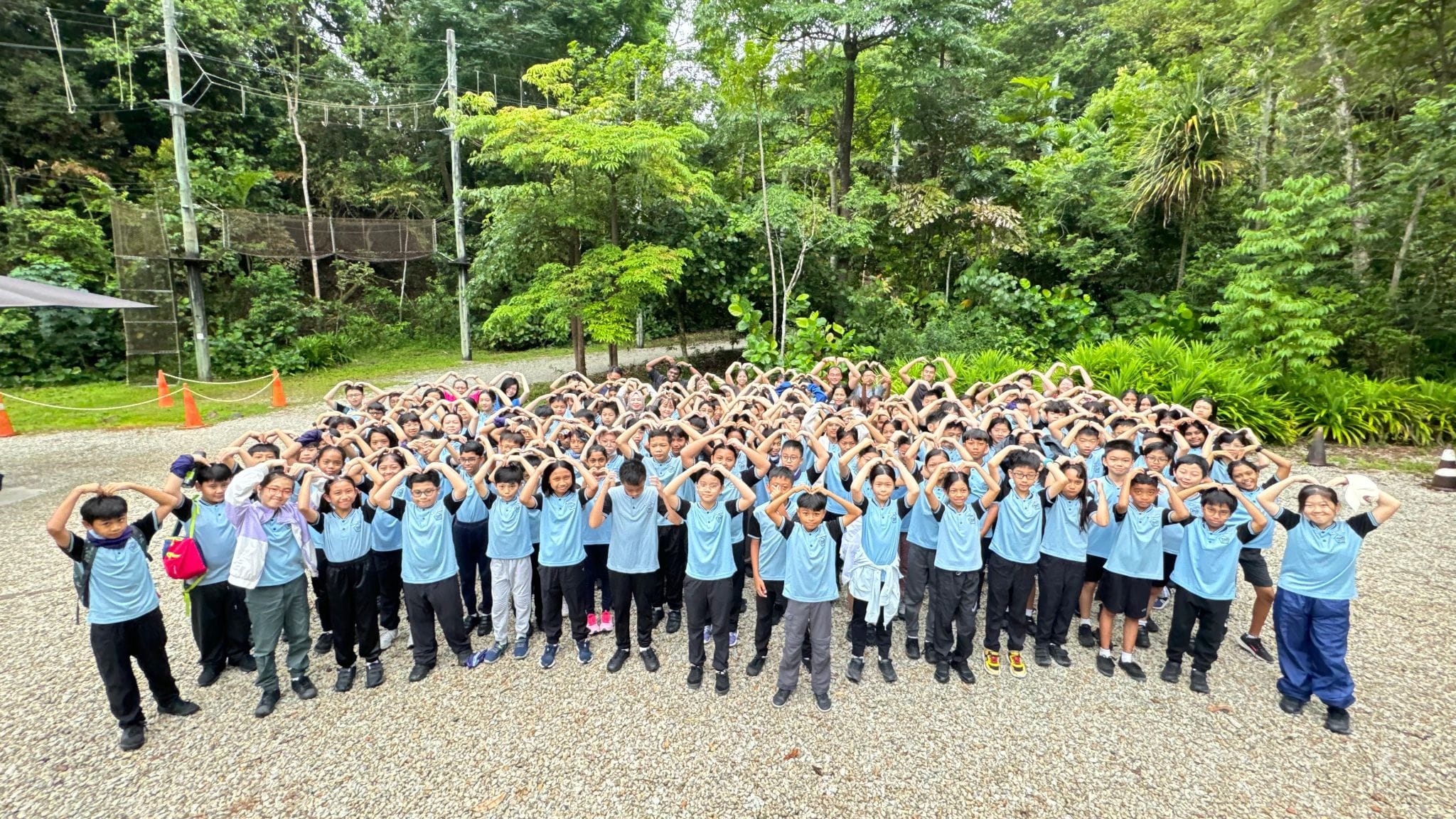 Large group of children in blue uniforms posing outdoors, making heart shapes with their hands above their heads.