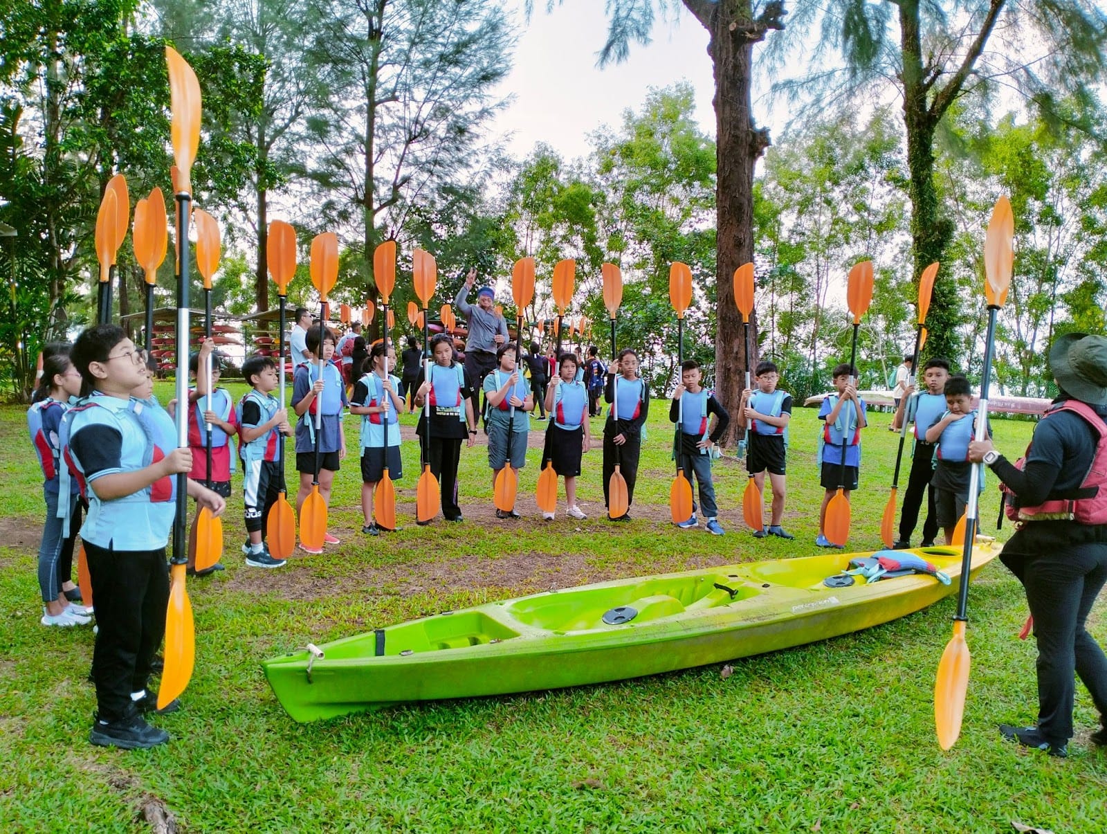 Children in life vests hold orange paddles around a green kayak on grass, receiving instruction outdoors near trees.