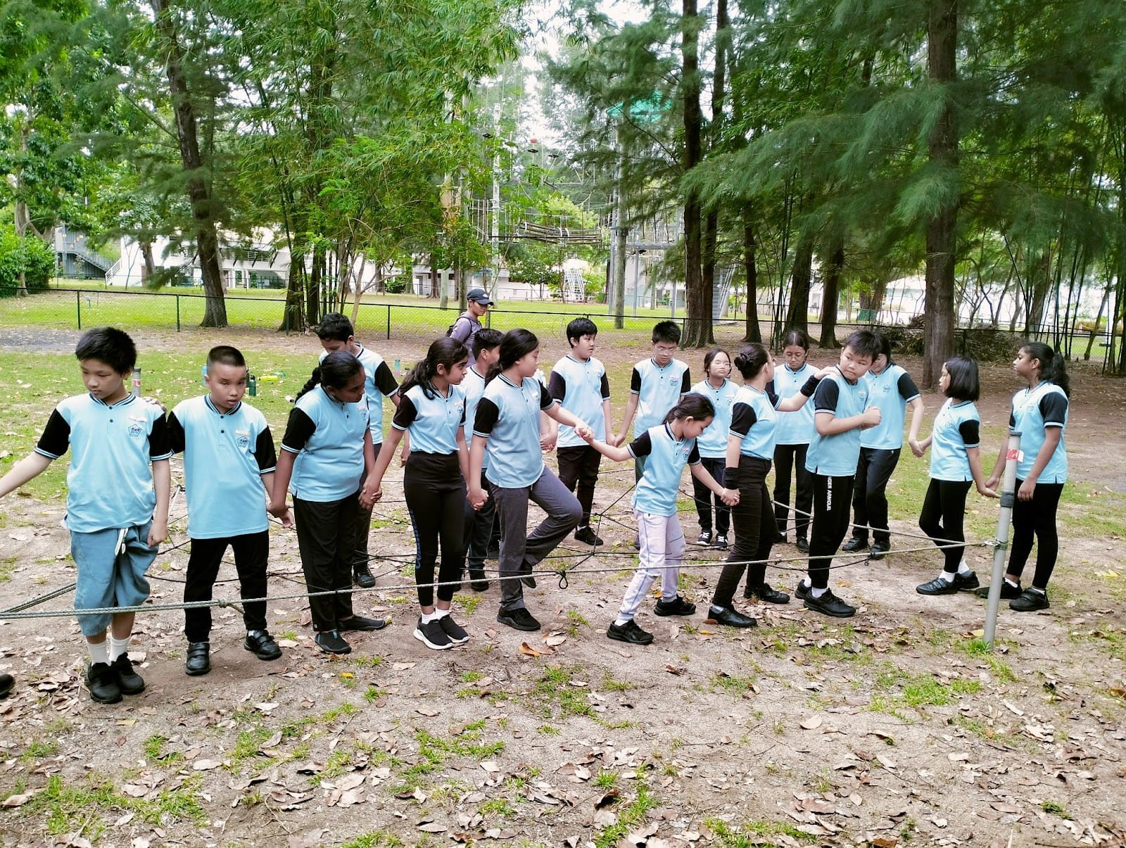 Students in light blue and black uniforms hold hands in a circle outdoors, navigating a rope obstacle course.