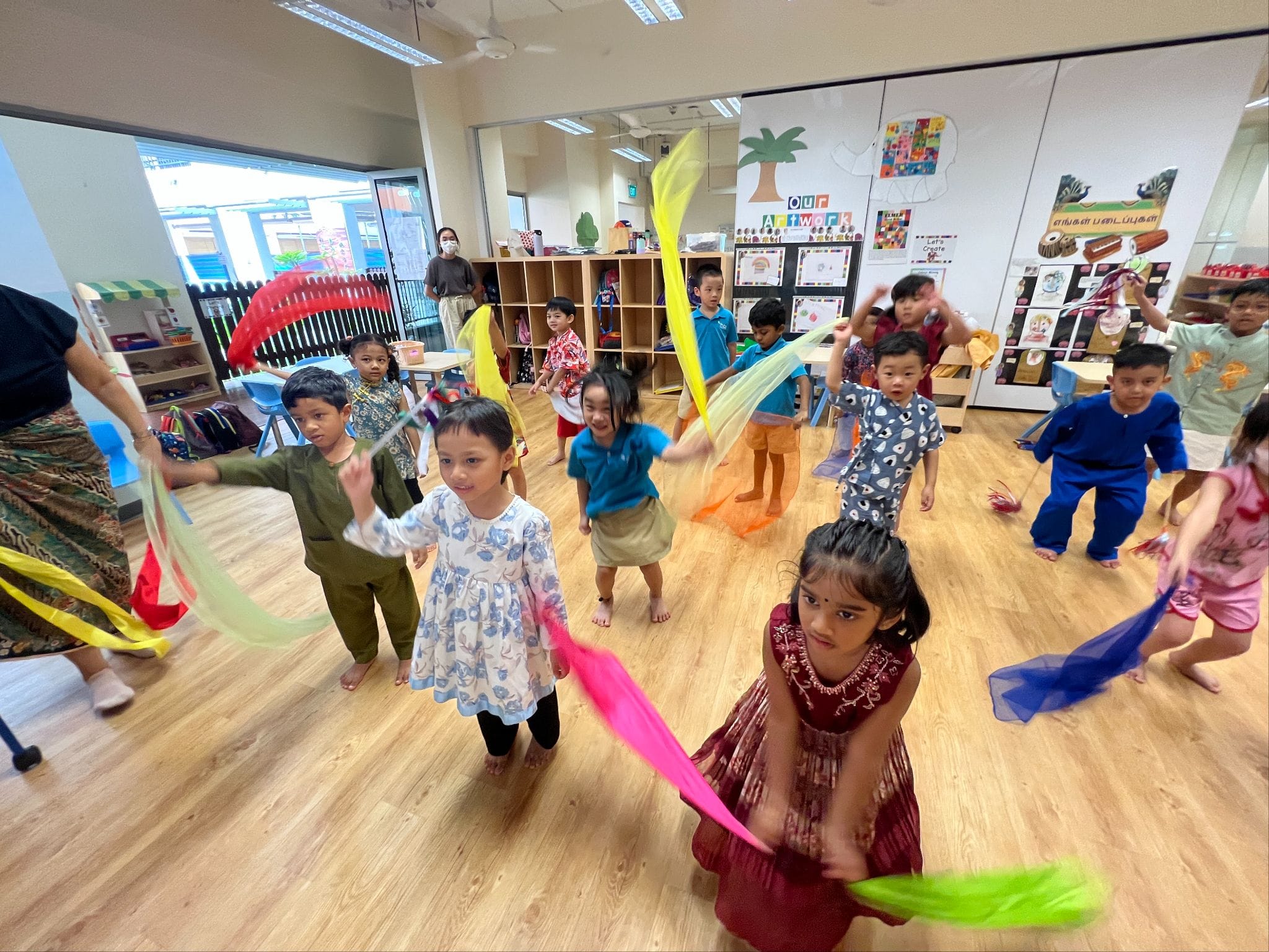 Young children wave colorful fabric scarves while dancing barefoot in a bright classroom supervised by adults.