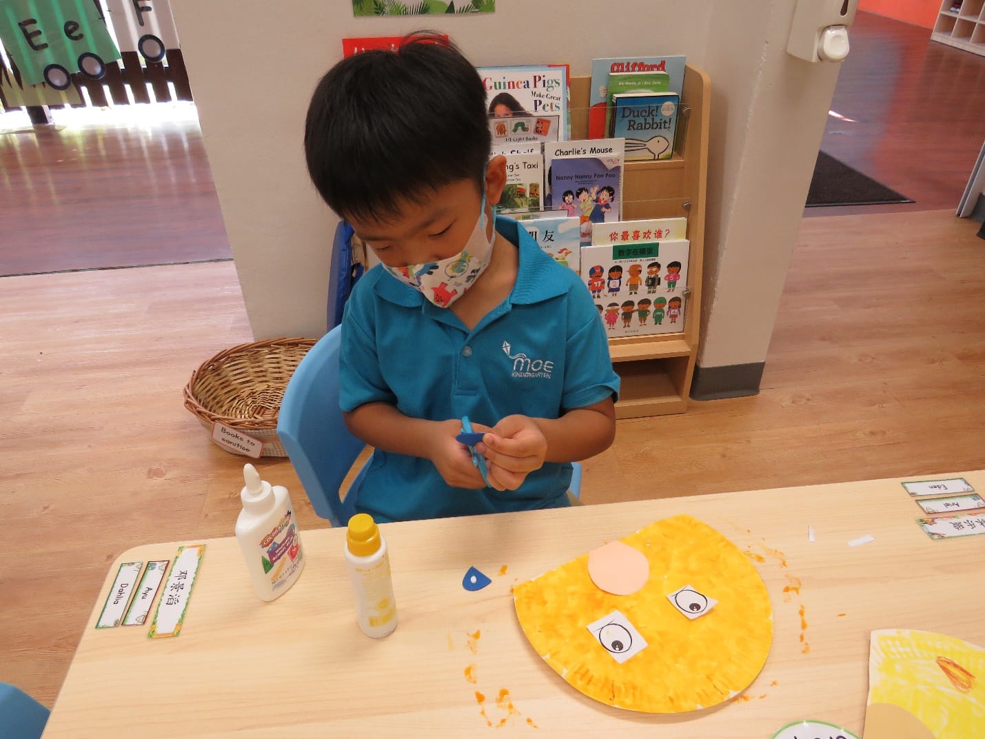 Young masked boy in blue uniform doing a craft project with a yellow paper plate face at a classroom table.