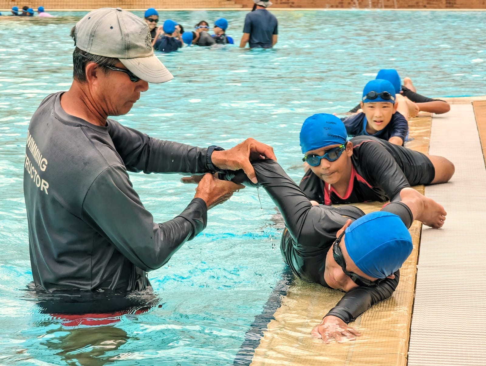 A swimming instructor in a pool guides children wearing blue caps and goggles along the pool edge during a lesson.