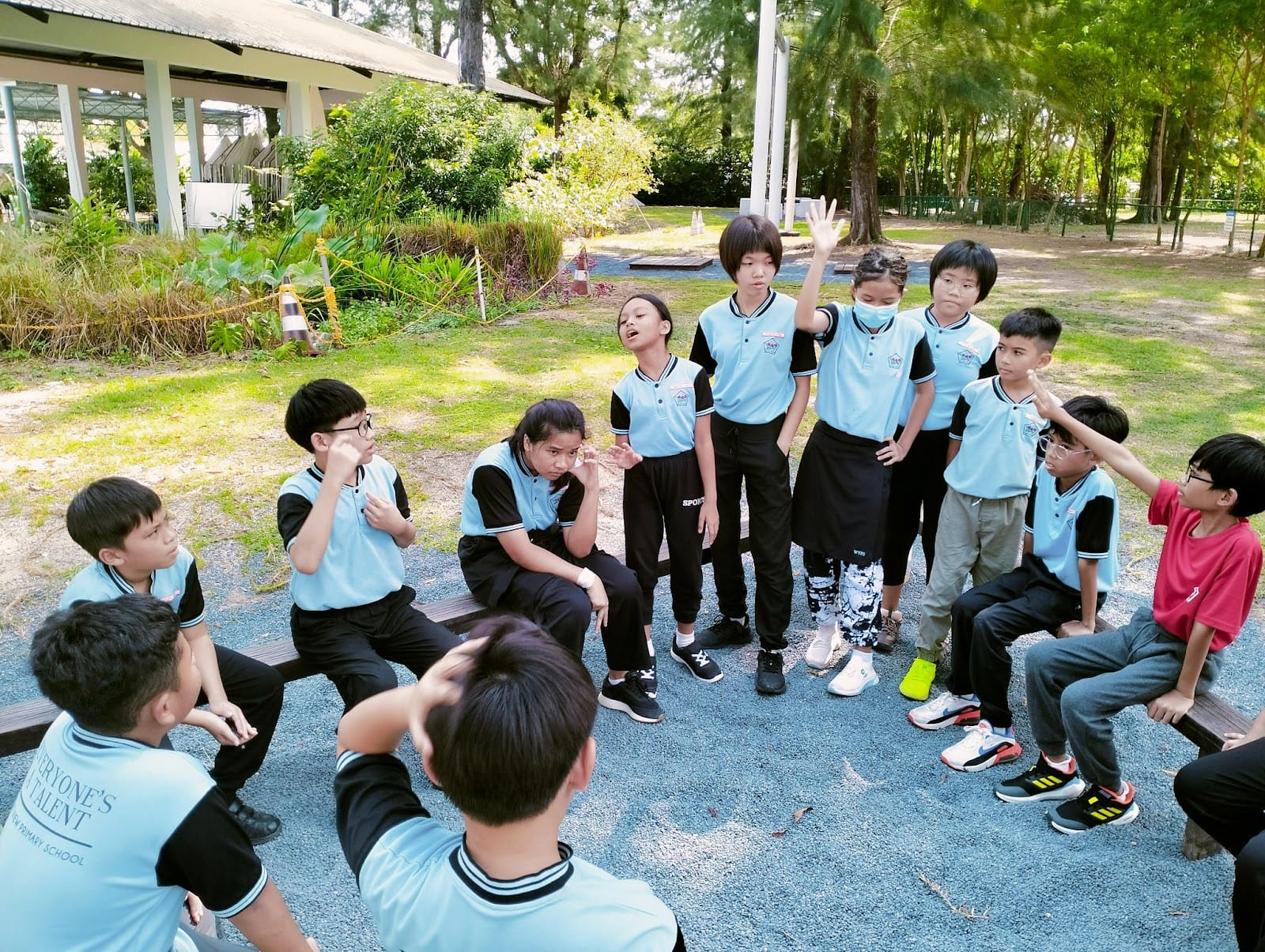 A group of primary school children in light blue uniforms gather outdoors, some standing and gesturing during a group activity.