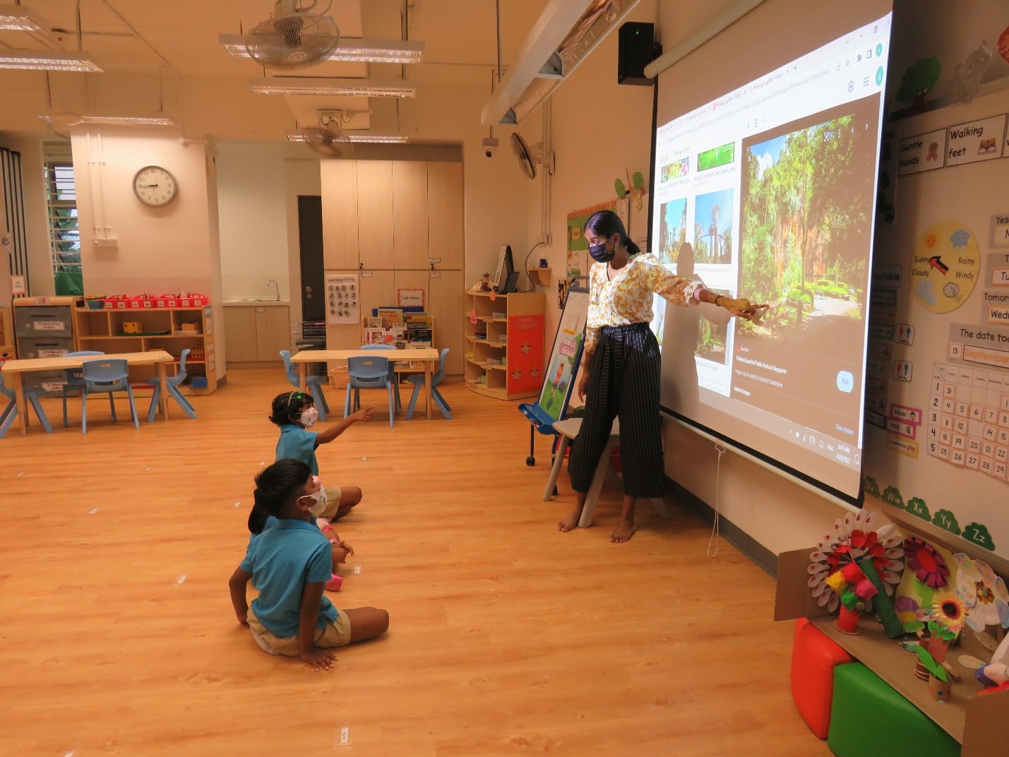 A masked teacher stands by a projector screen showing park images while two young children sit on the classroom floor.