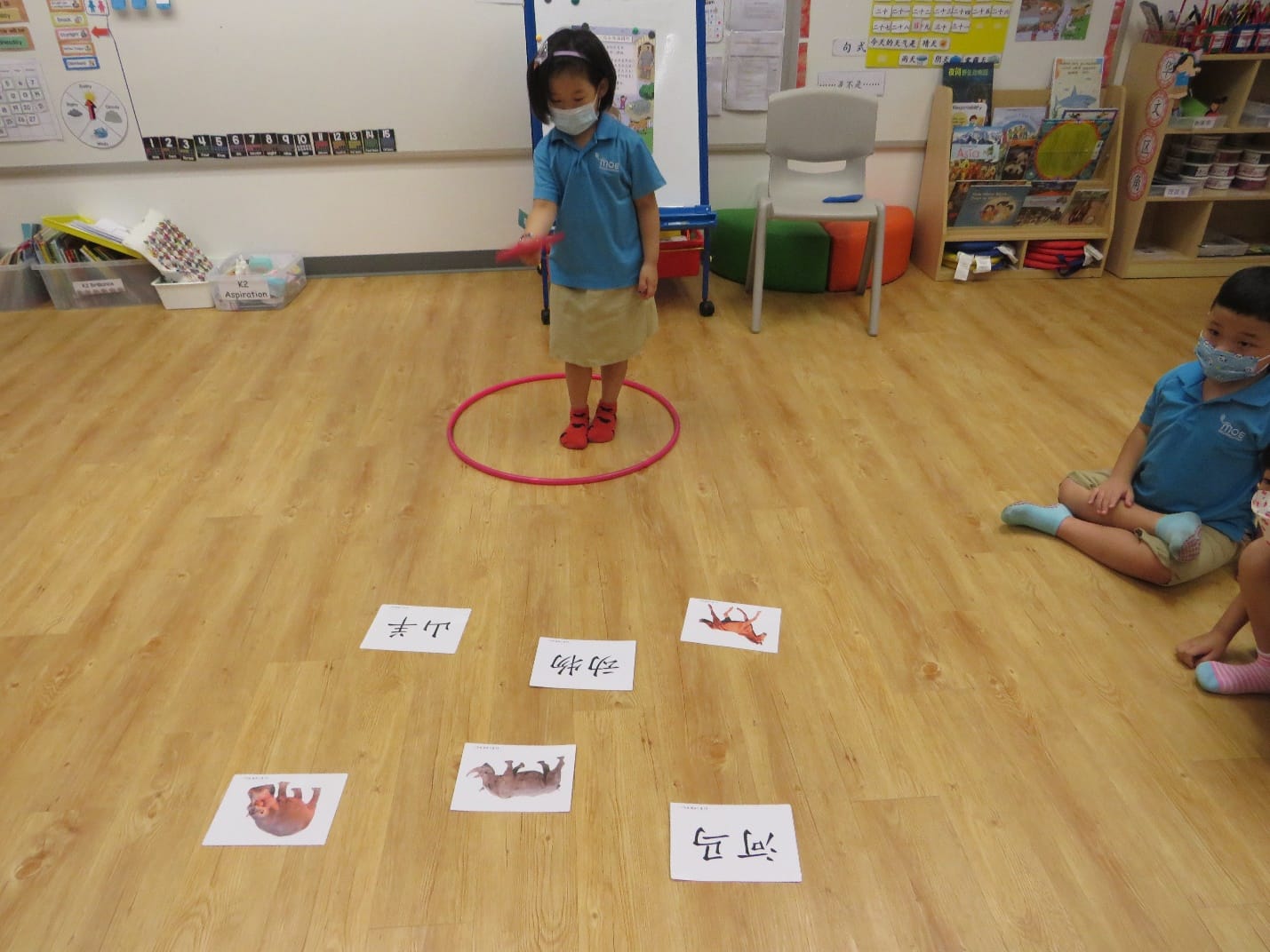 Young girl in a mask stands inside a pink hula hoop on a classroom floor with Chinese character cards around her.