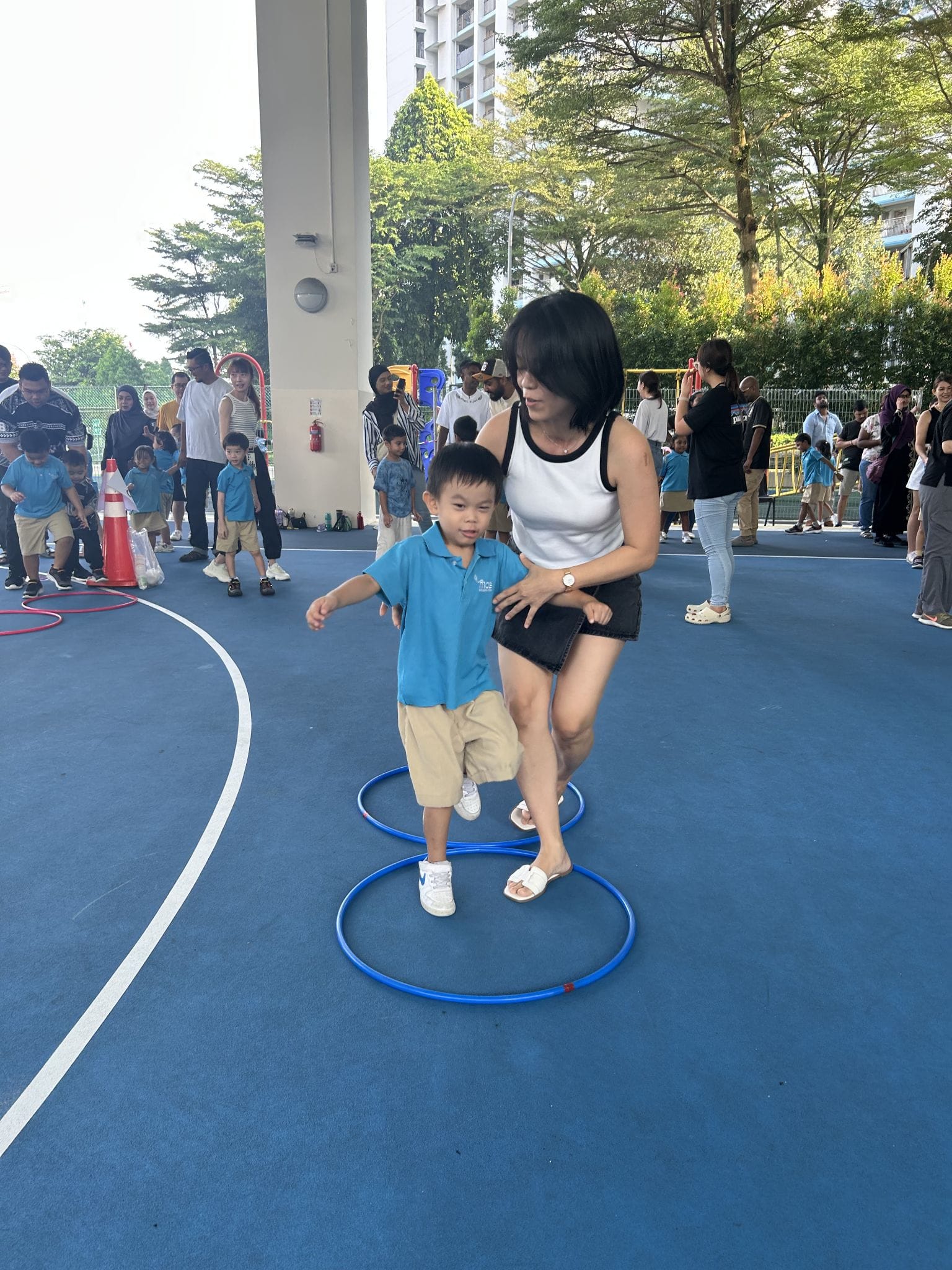 A woman guides a young boy stepping through a blue hula hoop on an outdoor court during a children's activity event.