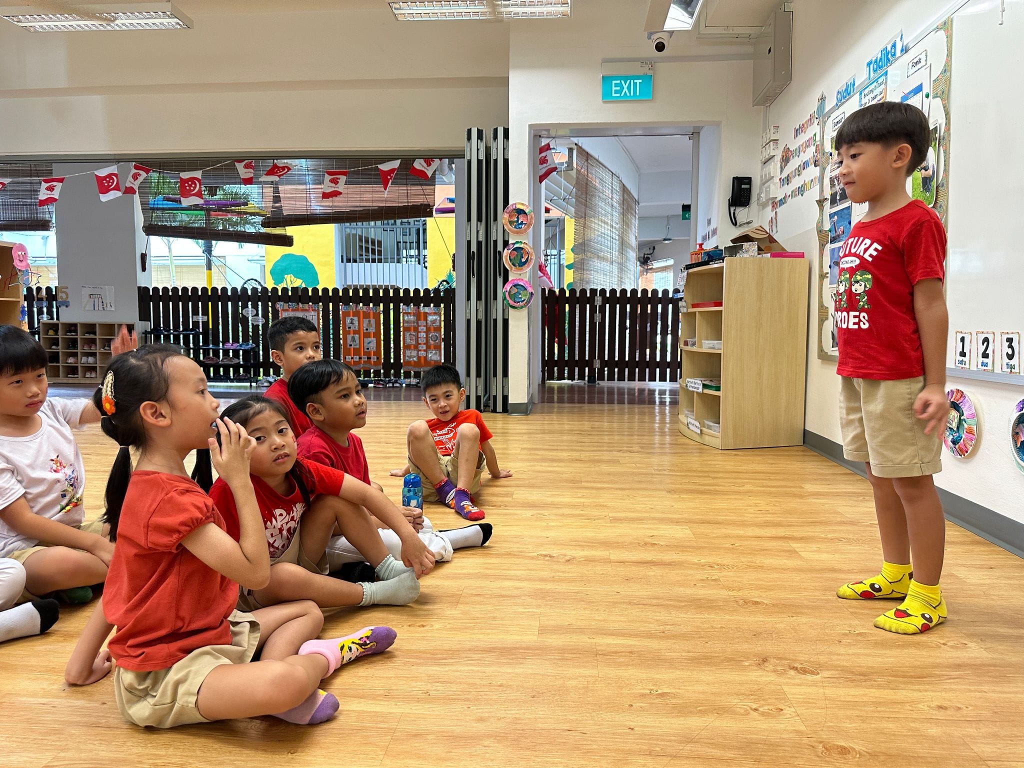 Young children sit on a classroom floor looking up at a boy in a red shirt standing before them.