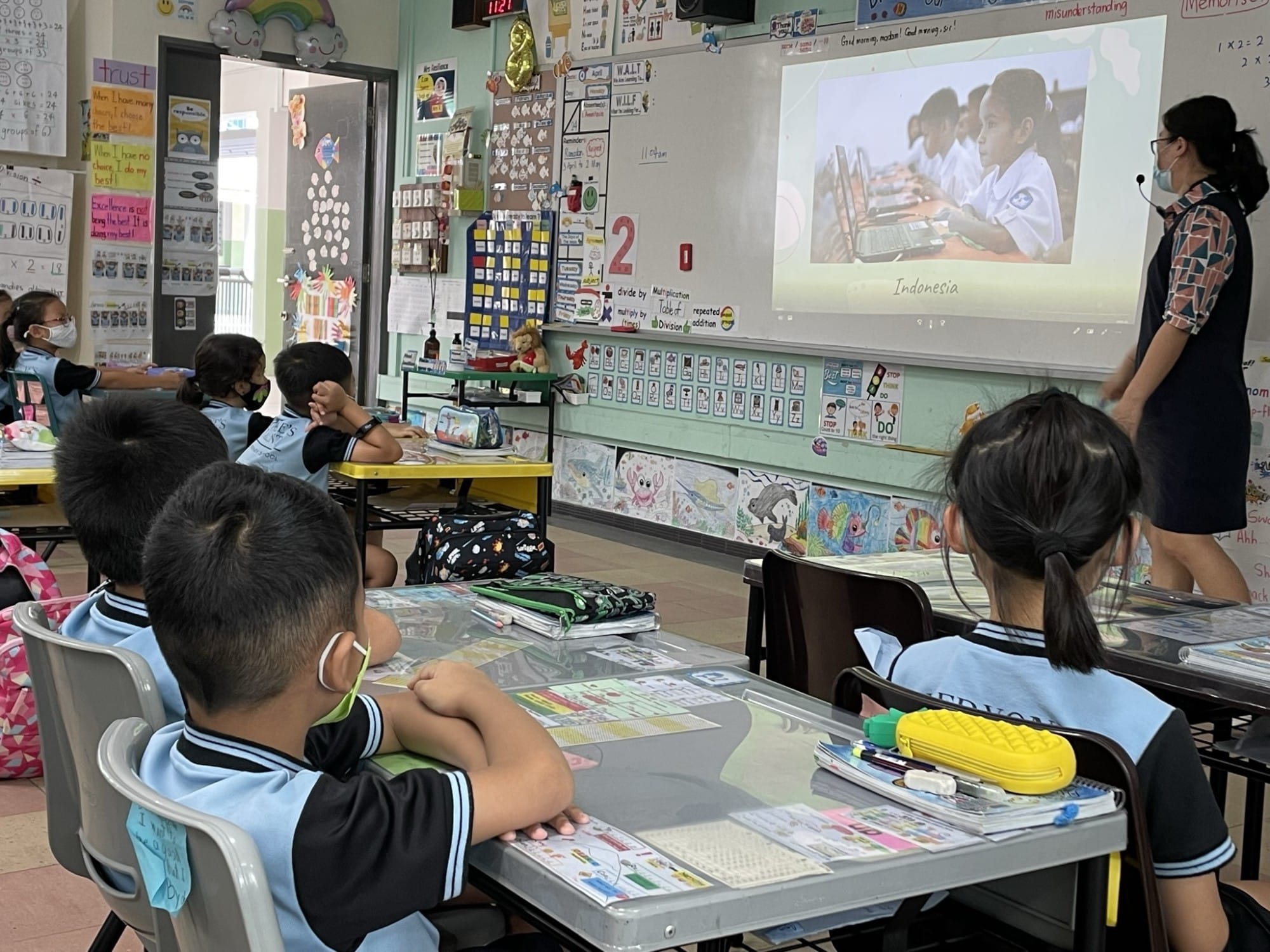 Young students in uniforms sit at desks watching a projected slide labeled Indonesia shown by a masked teacher.