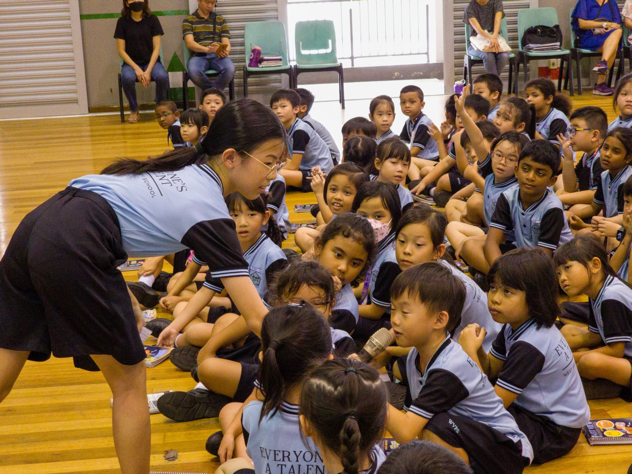A teacher in a blue uniform leans toward a large group of young students in matching uniforms seated on a gym floor.