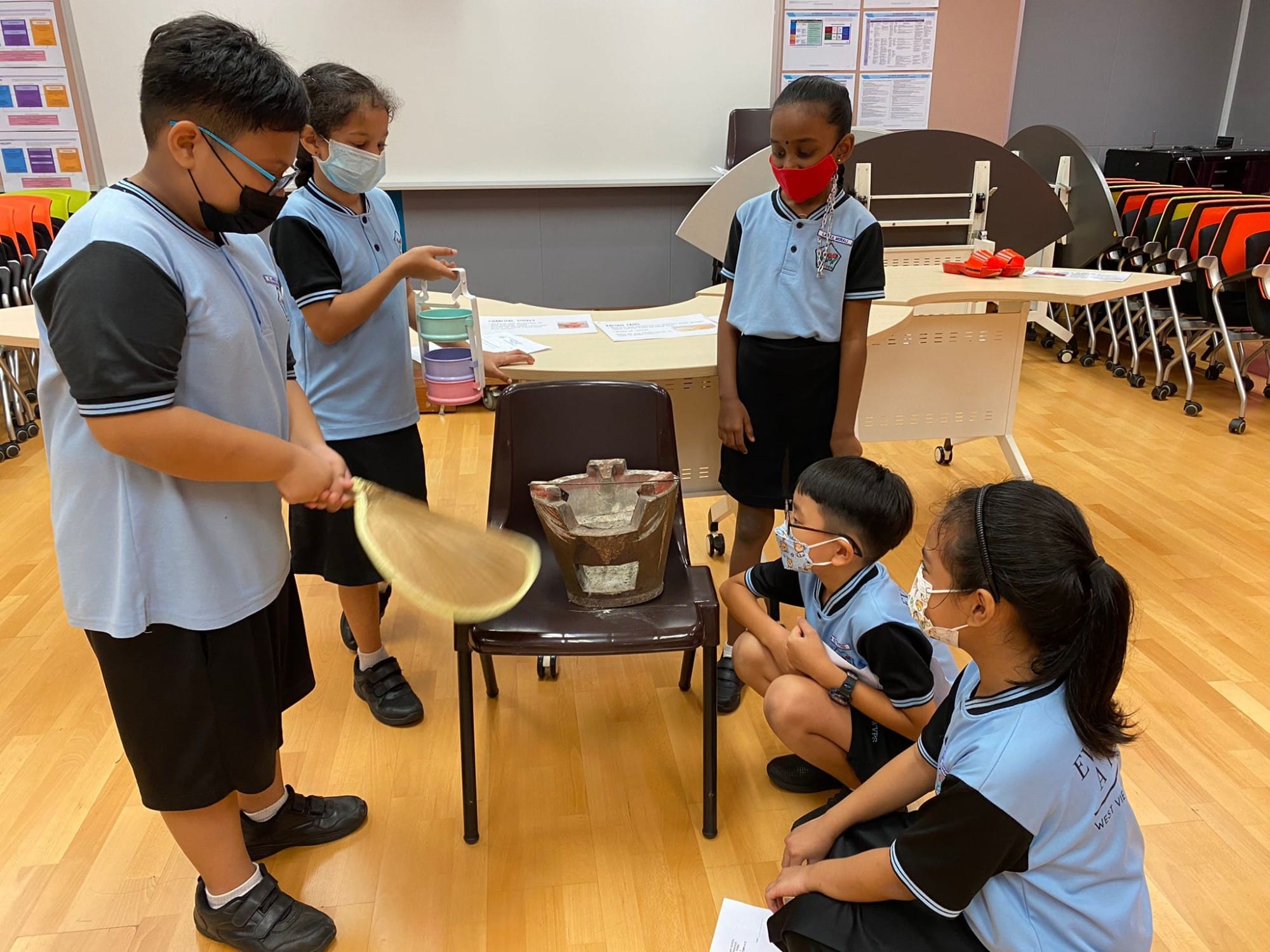 Five masked students in blue school uniforms gather around a chair holding a bucket during a classroom activity.