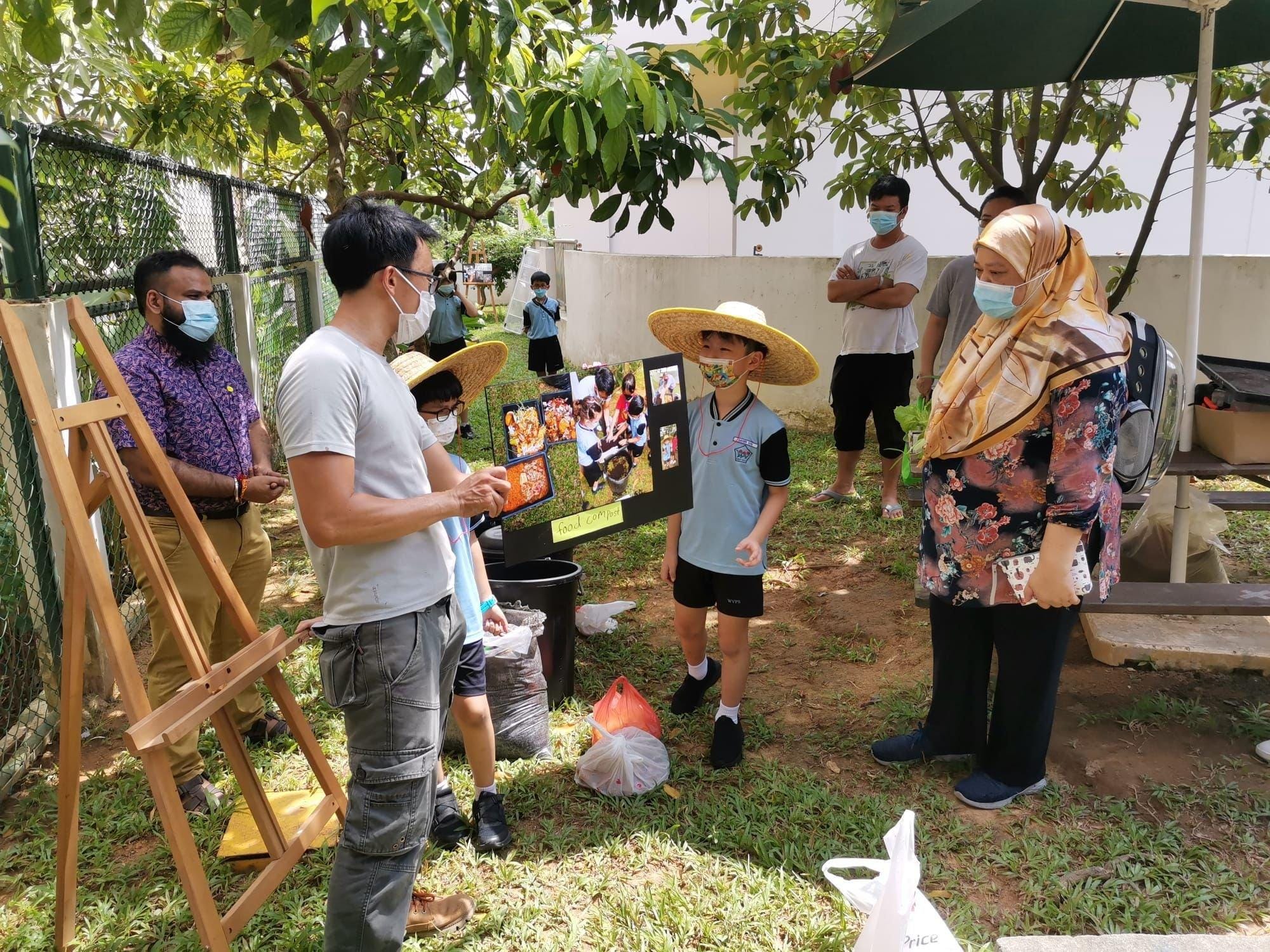 A masked child in a straw hat presents a food compost poster board to masked adults outdoors in a garden.
