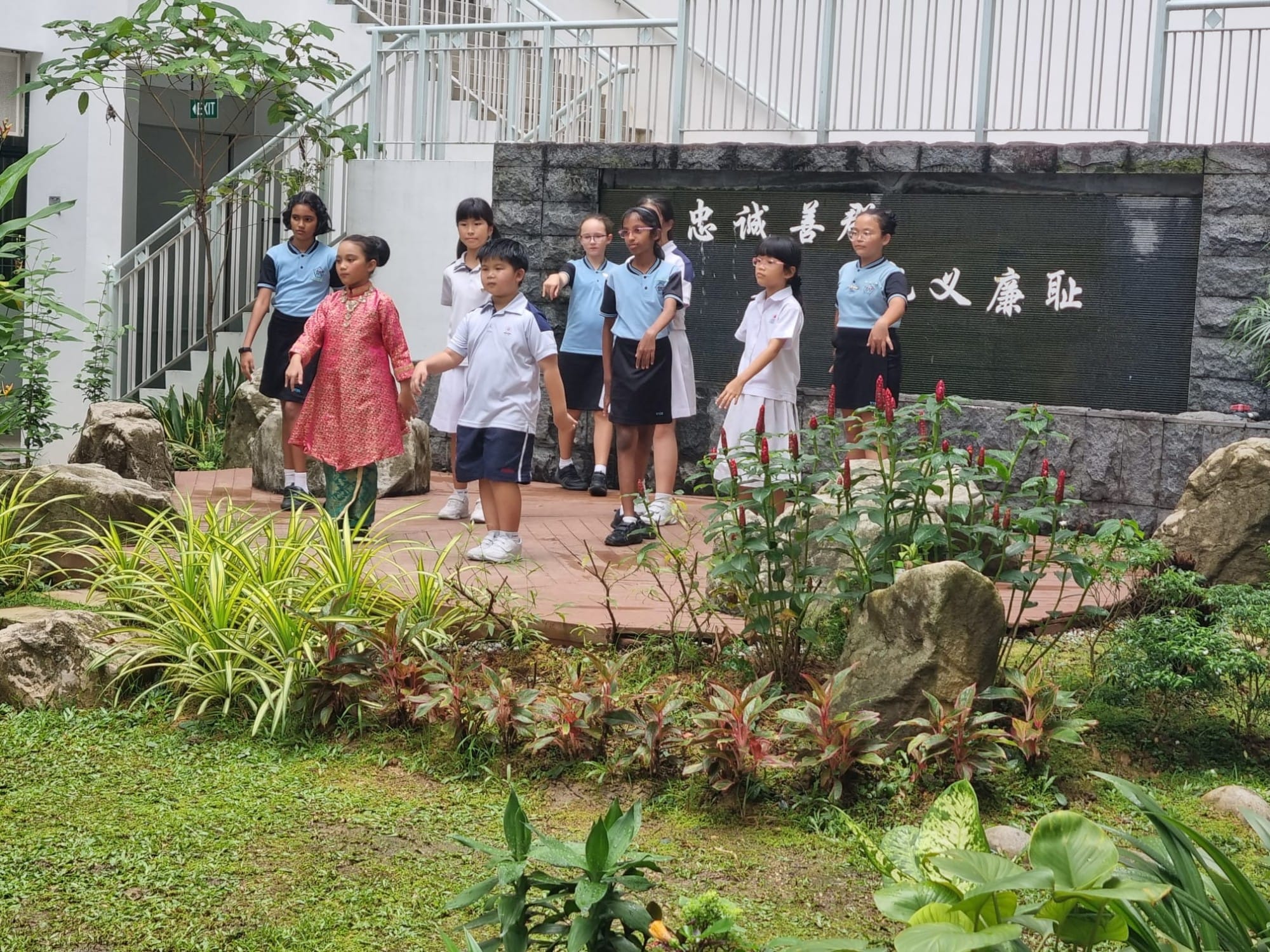 A group of schoolchildren in uniforms stand on a garden path in front of a stone wall with Chinese characters.