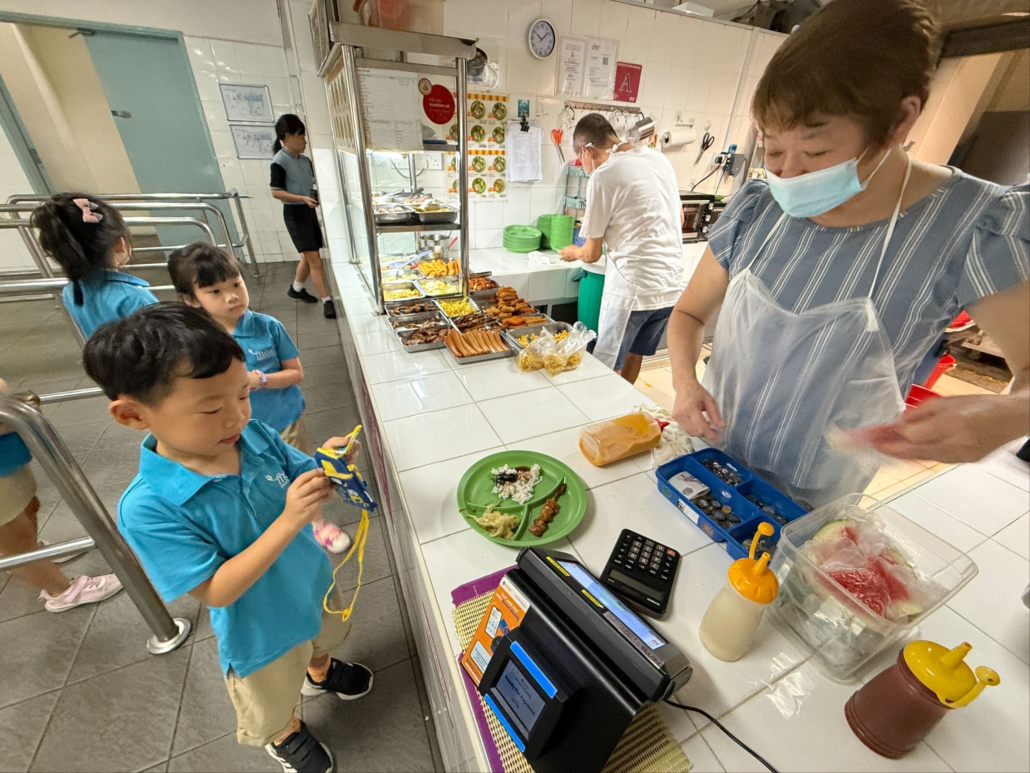 Young children in blue uniforms queue at a school canteen counter where a masked woman serves food.