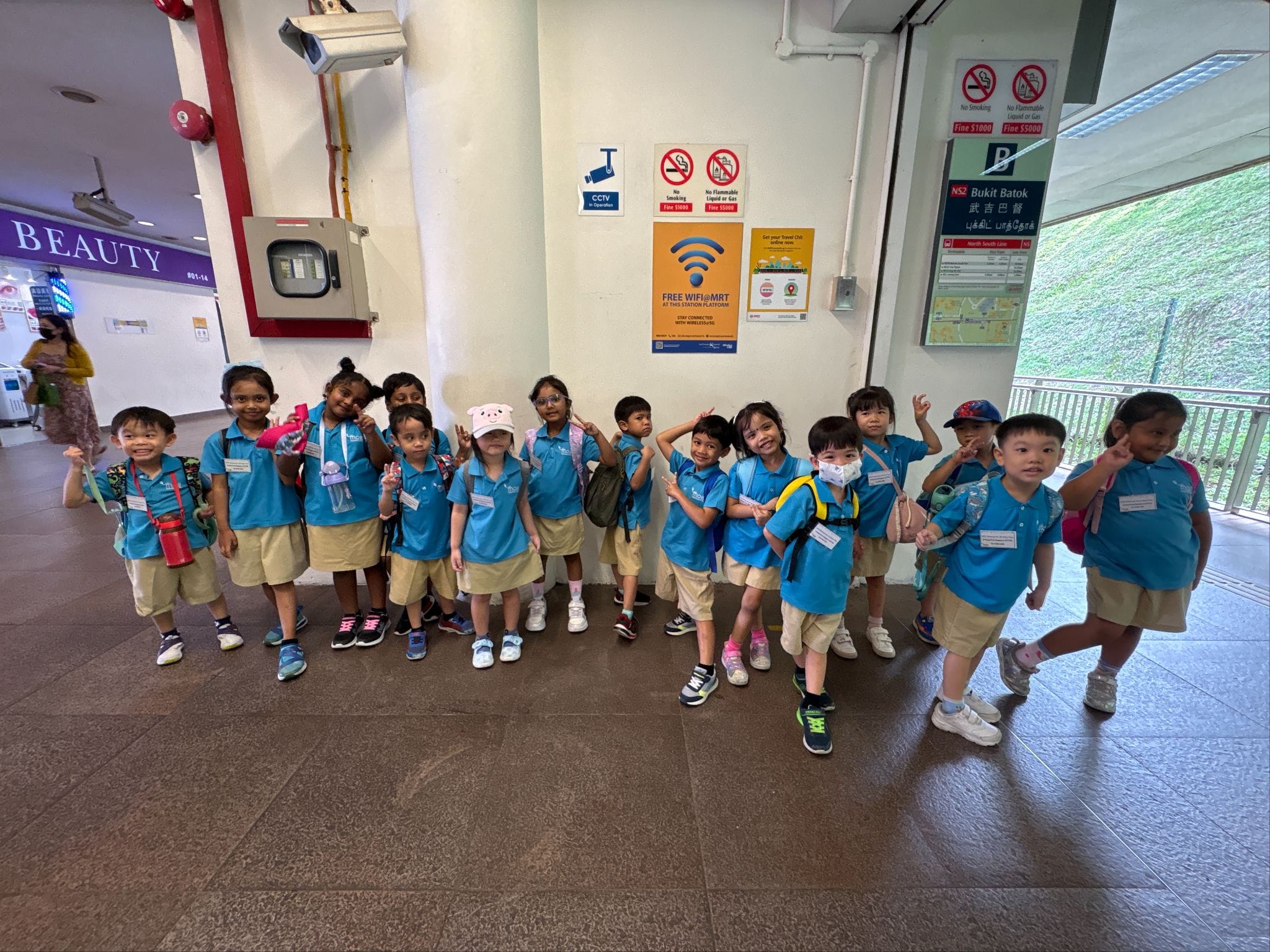 A group of young children in blue uniforms pose and smile at Bukit Batok MRT station in Singapore.