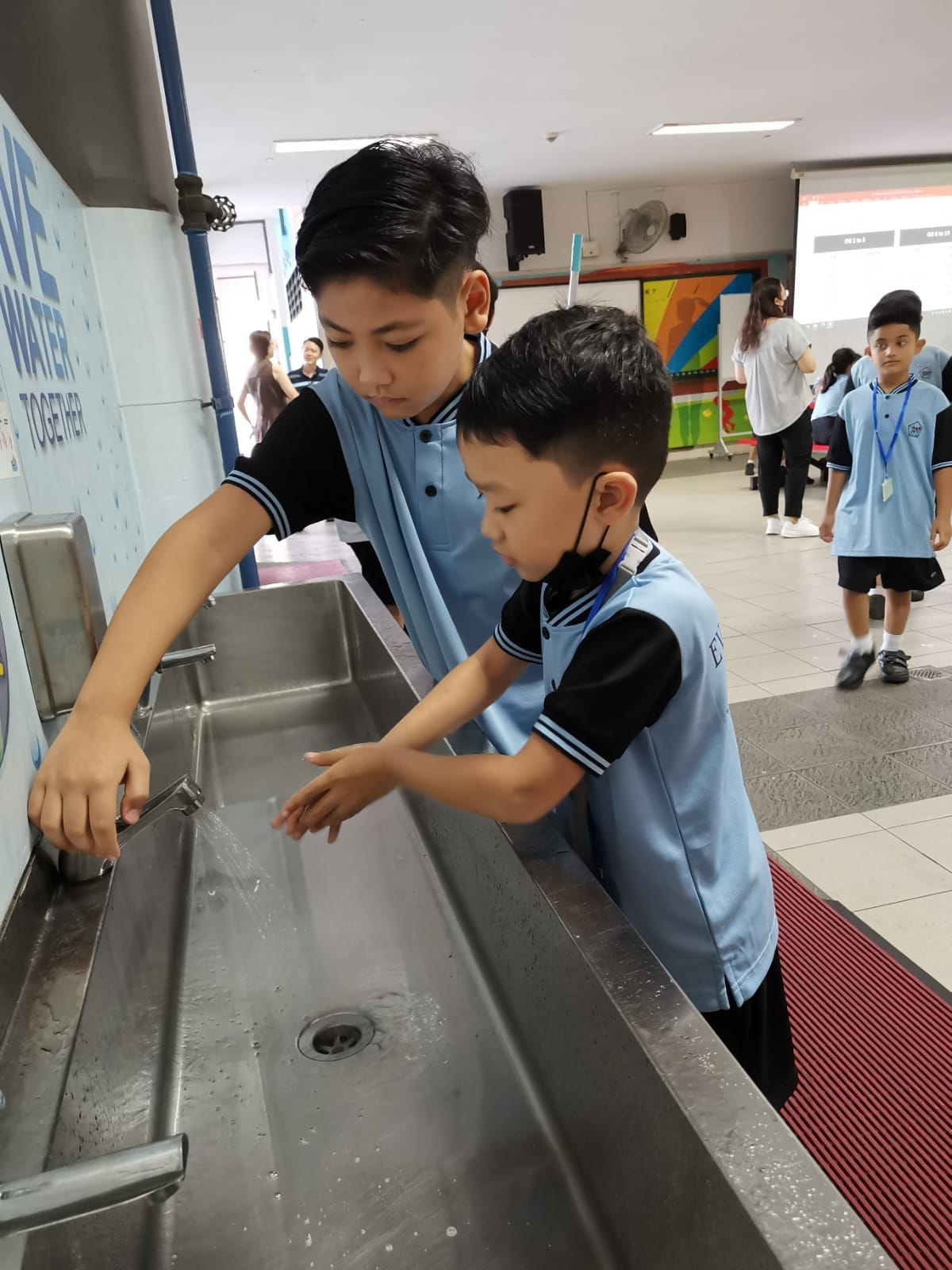 Two boys in blue school uniforms wash hands at a stainless steel sink in a school hallway.