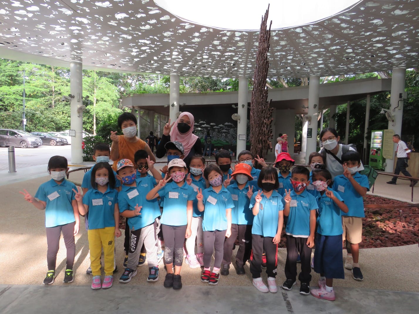 Group of young children in blue shirts and face masks pose with peace signs under a decorative pavilion outdoors.