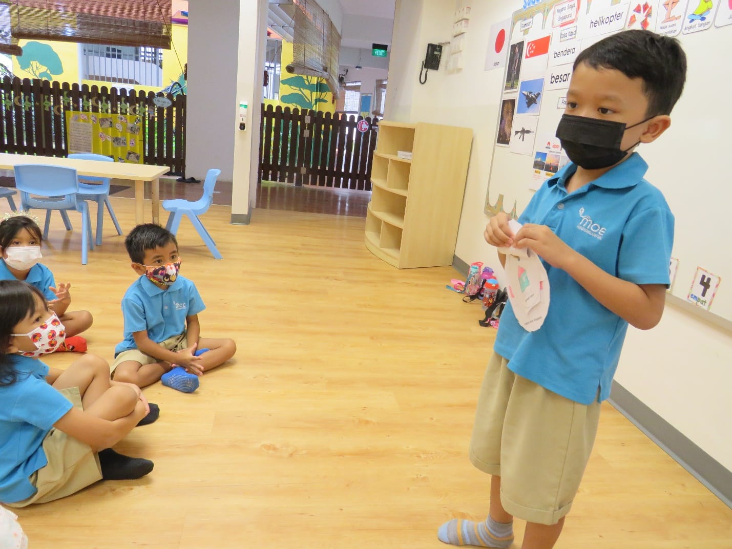 A masked boy in a blue school uniform stands holding paper while classmates sit on a classroom floor.