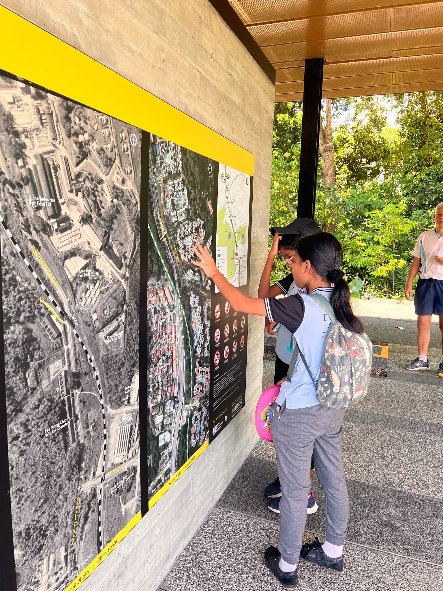 Two children with backpacks point at large outdoor map boards mounted on a wall near green trees.