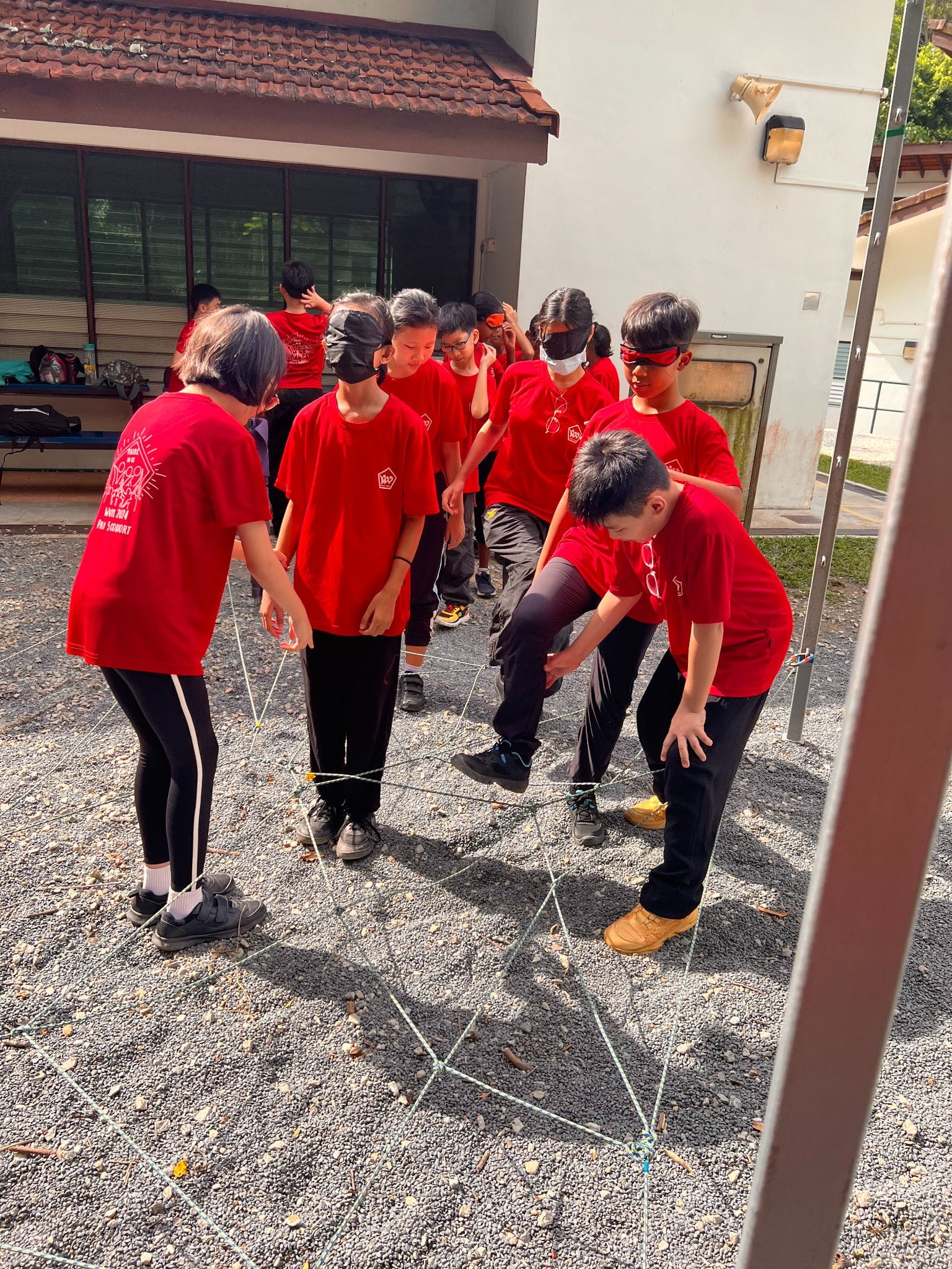 Children in red shirts, some blindfolded, navigate a string obstacle course on gravel outdoors.