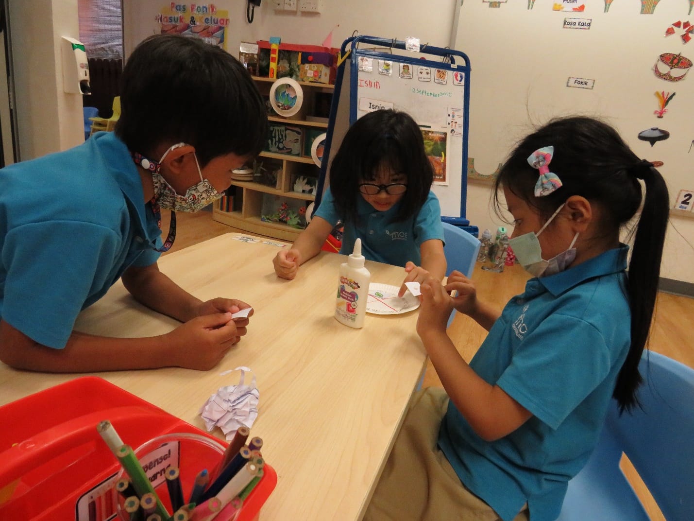 Three young students in blue uniforms and face masks do a paper craft activity at a classroom table with glue.