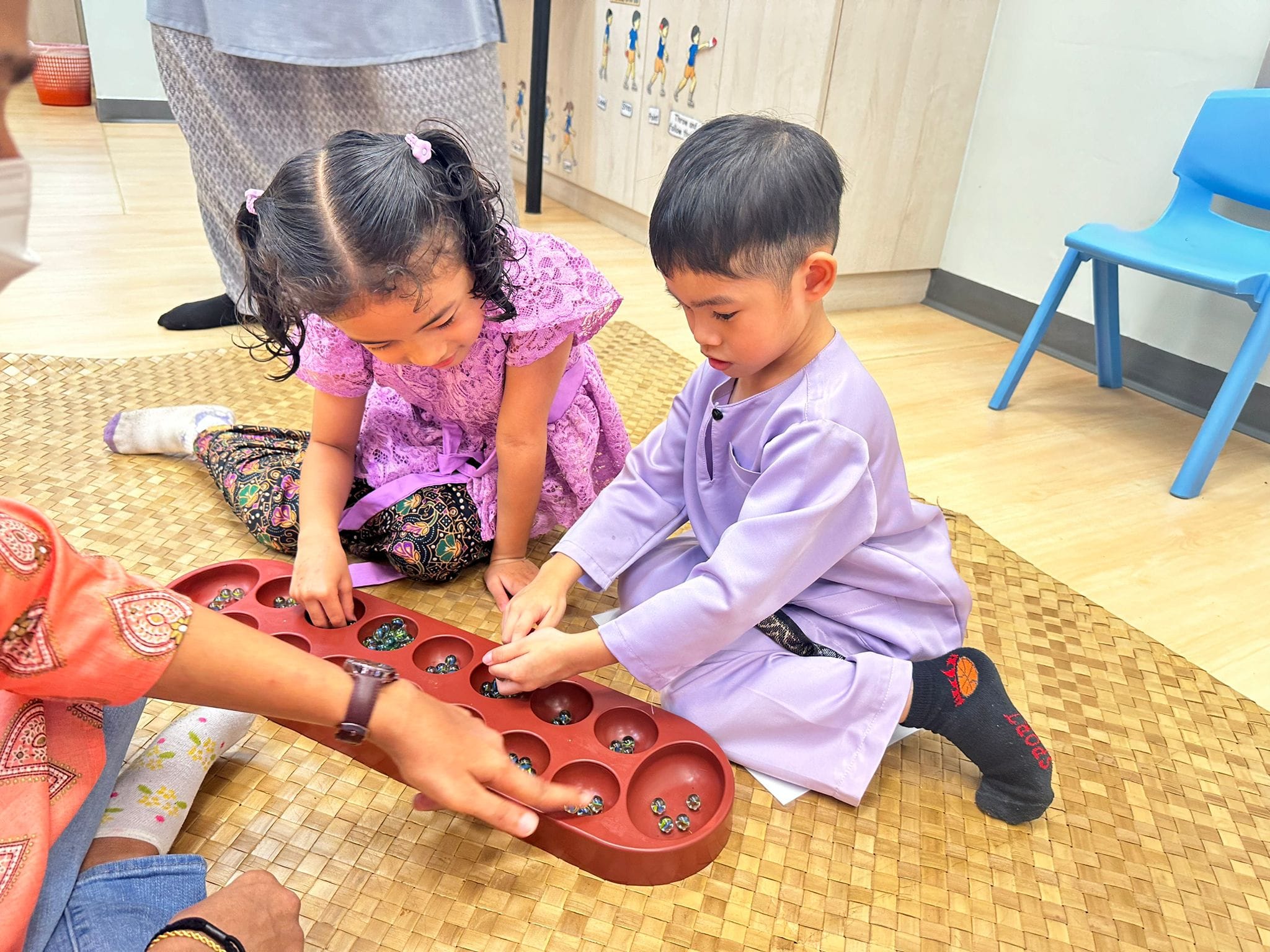 Two young children in traditional attire play a mancala board game with marbles on a woven mat indoors.