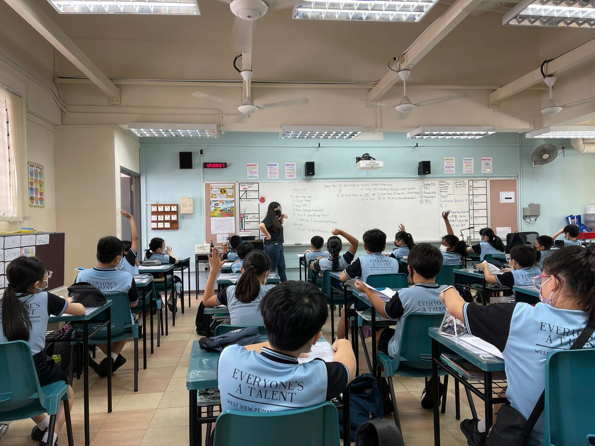 Students in light blue uniforms sit at desks facing a teacher at a whiteboard, with several raising their hands.