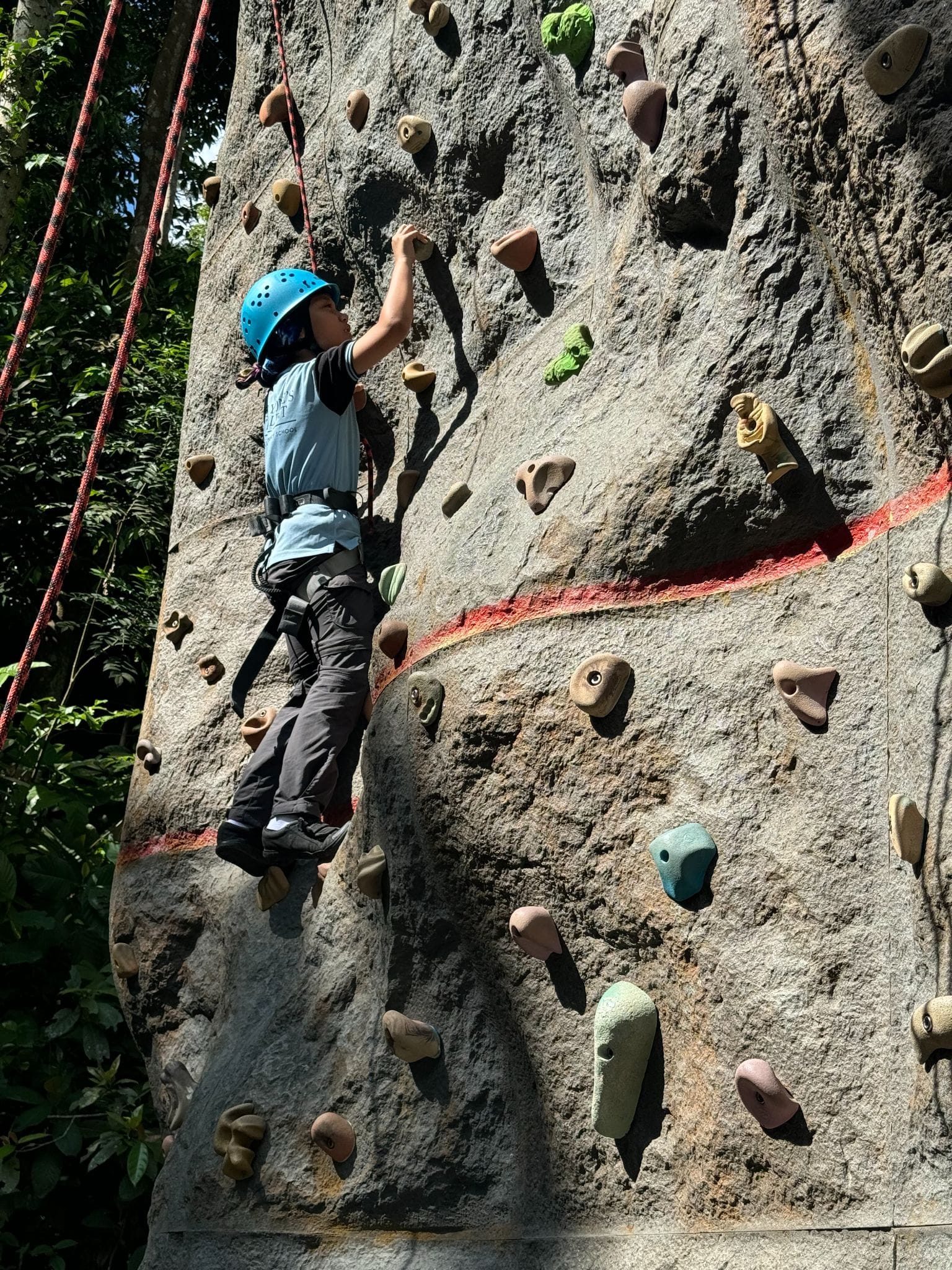 Child wearing a blue helmet and harness climbing an outdoor rock wall with colorful holds and red rope.