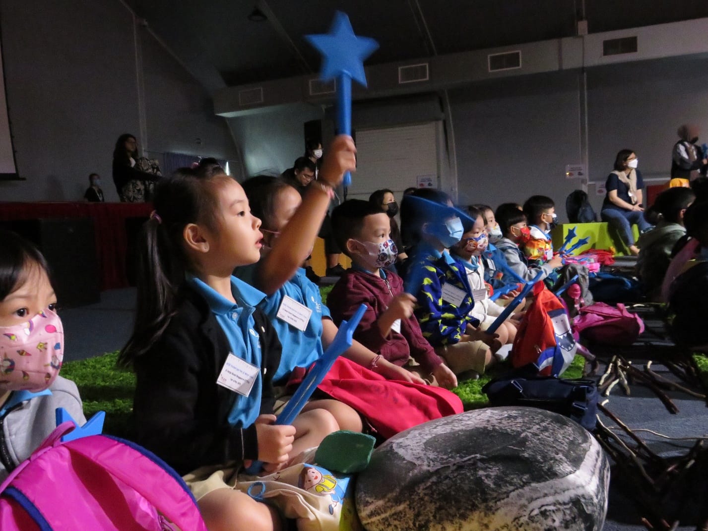 Children wearing masks sit in a row at an event, holding blue star-topped wands, with backpacks nearby.