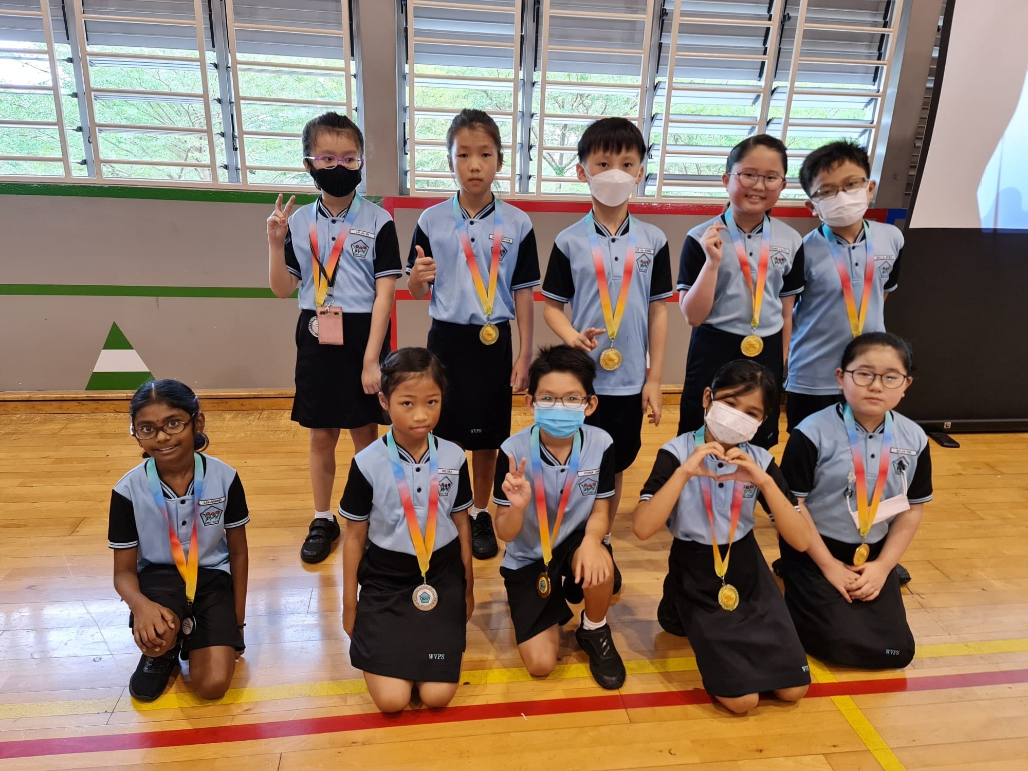 Ten young students in blue and black uniforms pose with gold medals on colorful ribbons in a school hall.