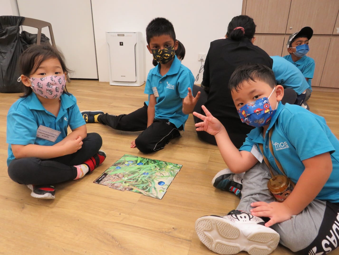 Three young children in teal uniforms and face masks sit on a floor around a nature picture card.