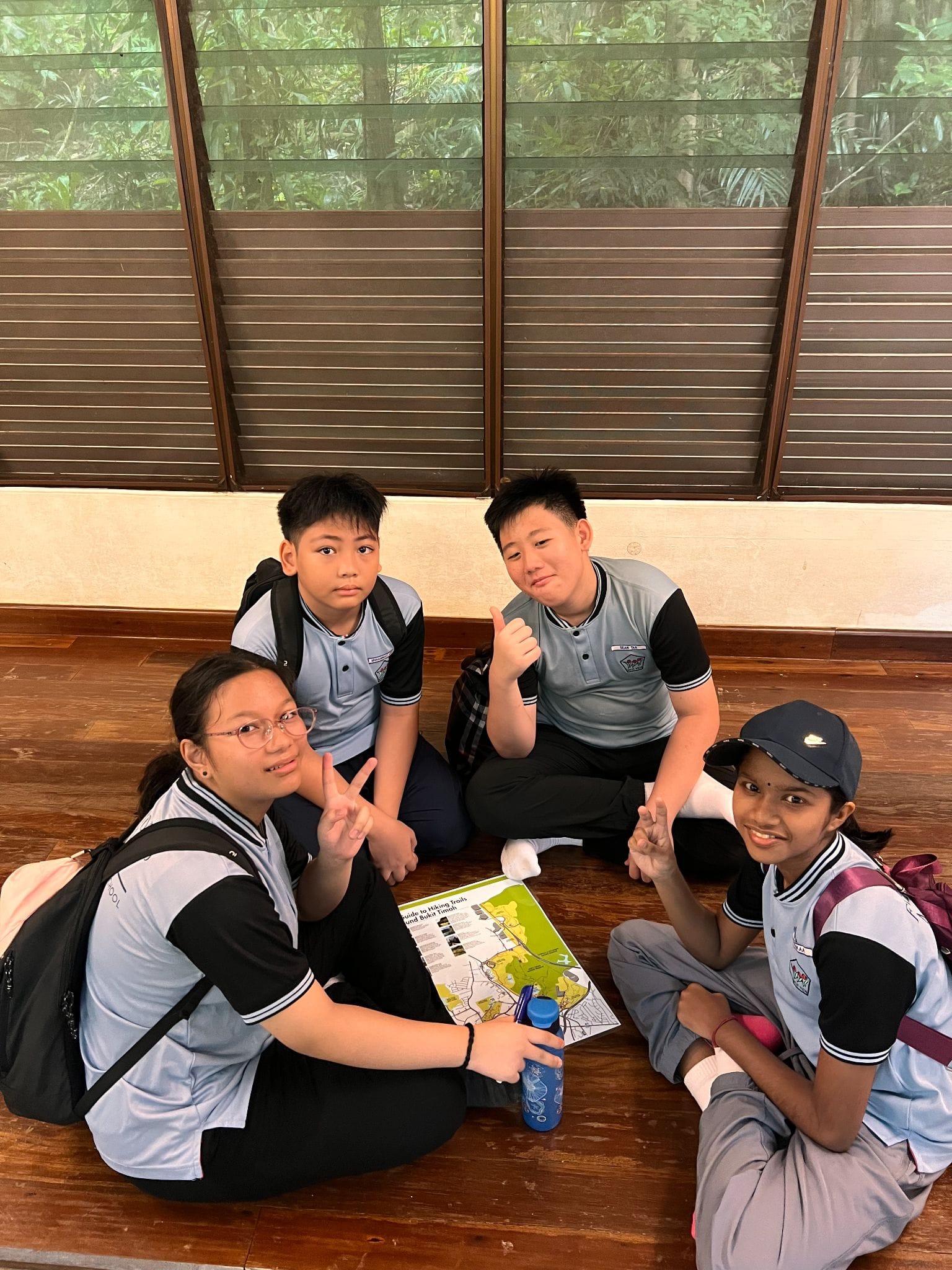 Four students in matching uniforms sit on a wooden floor, posing with peace signs and a thumbs up, holding a trail map.