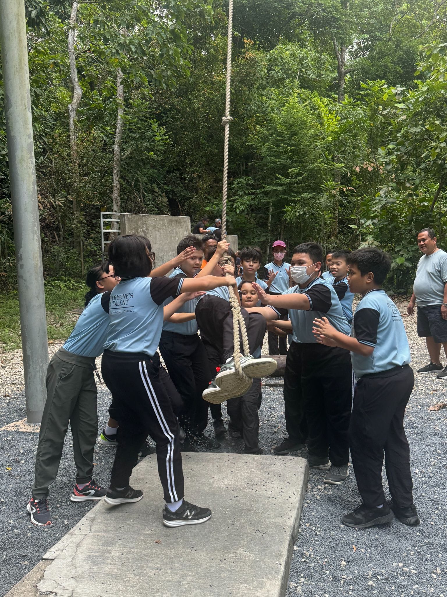 Students in light blue uniforms help a peer climb a hanging rope outdoors, surrounded by trees.