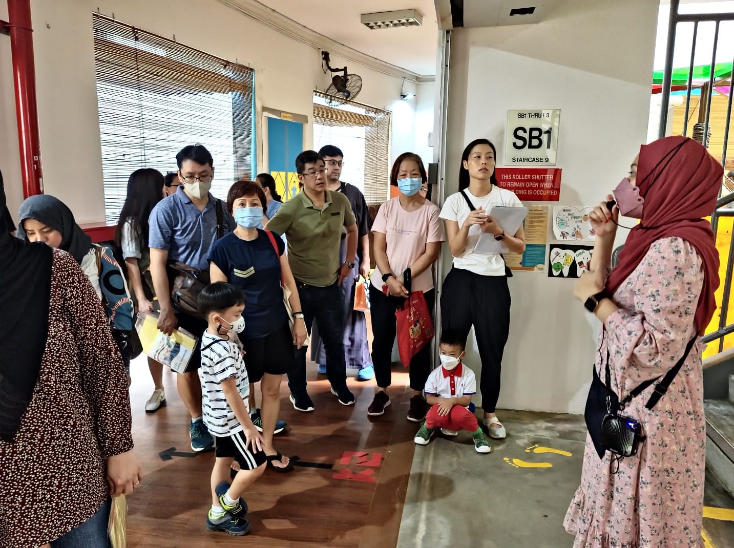 A group of adults and children wearing face masks gather in a corridor near a staircase sign labeled SB1.