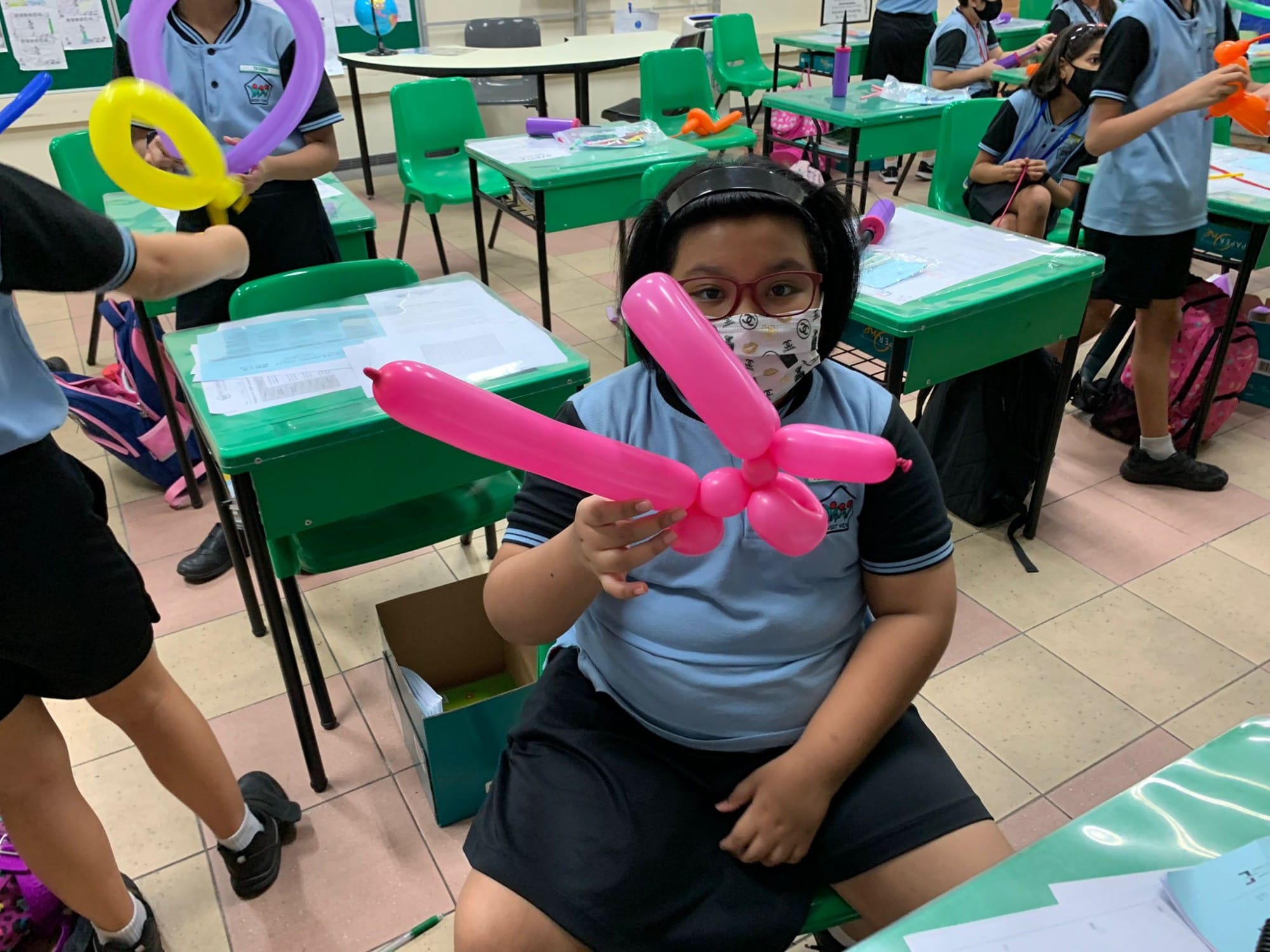 A masked schoolgirl holds a pink twisted balloon animal in a classroom with green desks and other students.