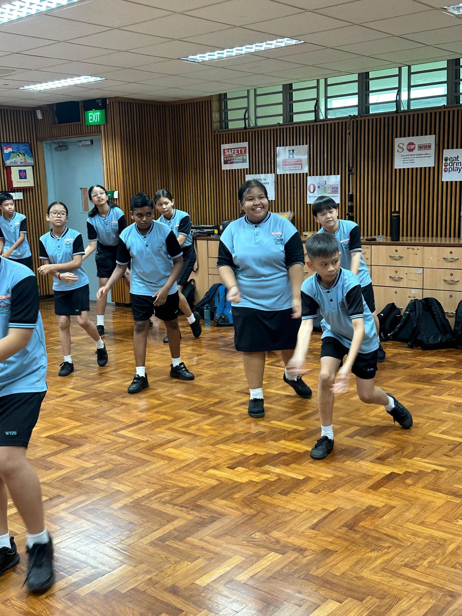 Students in matching blue and black uniforms dancing on a wooden parquet floor in a school hall.