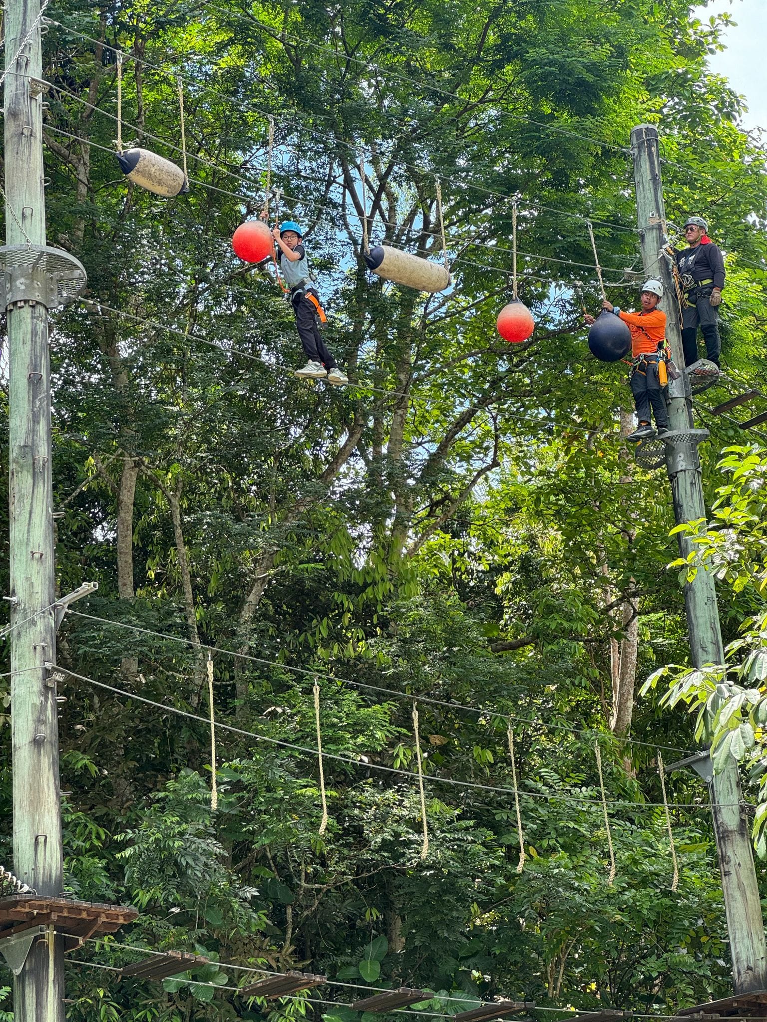 A child in a helmet navigates a high ropes course with hanging obstacles while two instructors watch from a pole.
