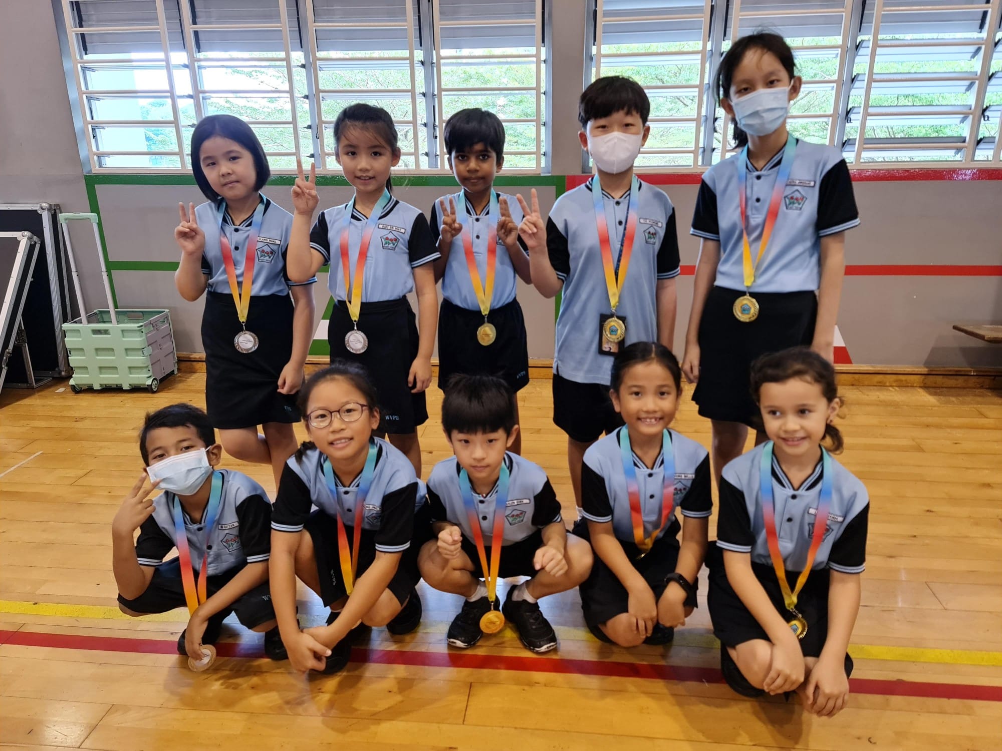 Nine young students in blue uniforms pose with medals on colorful ribbons, some making peace signs, in a gym.