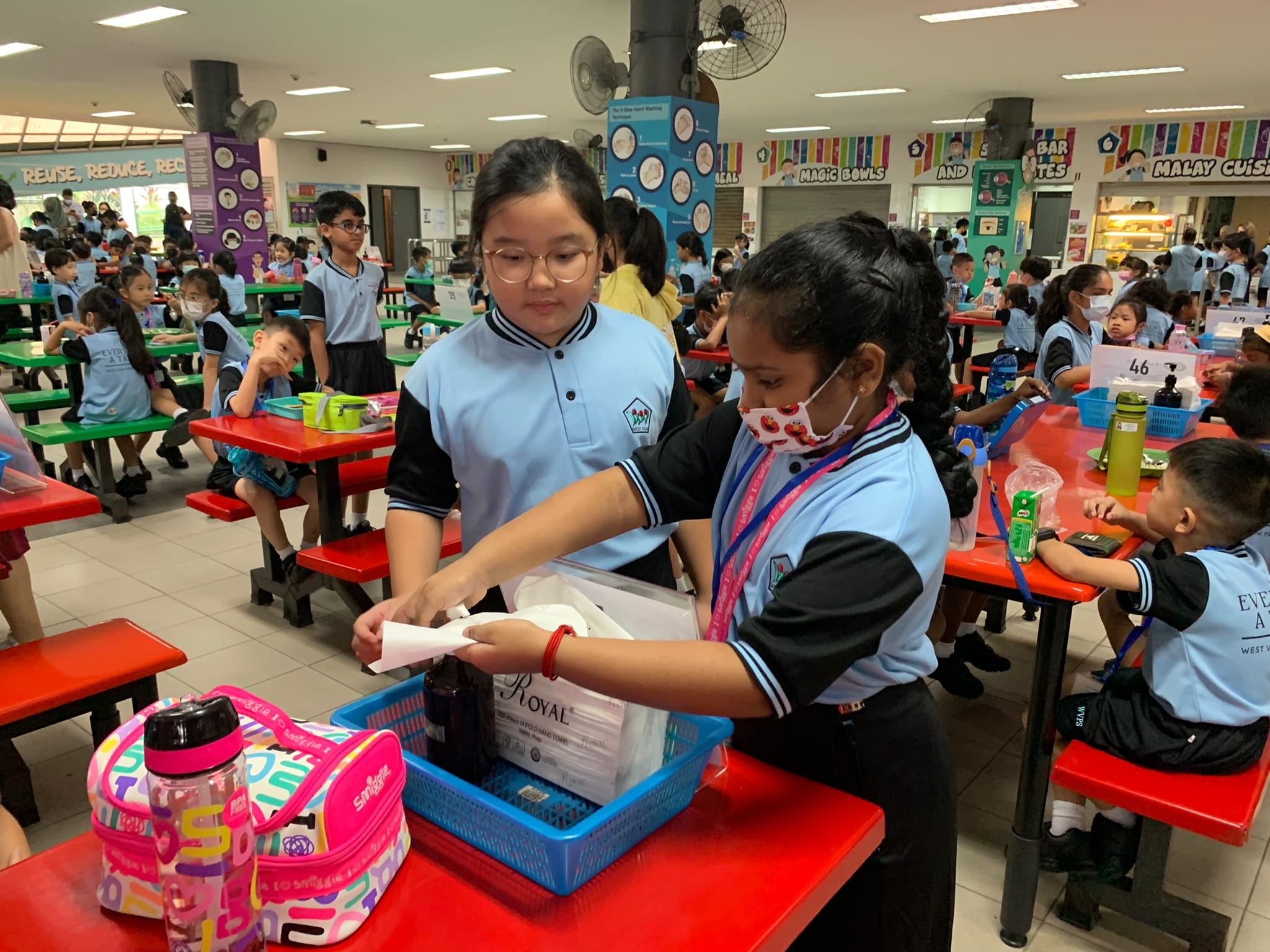 Two school girls in blue uniforms sort items into a blue basket in a busy school canteen.