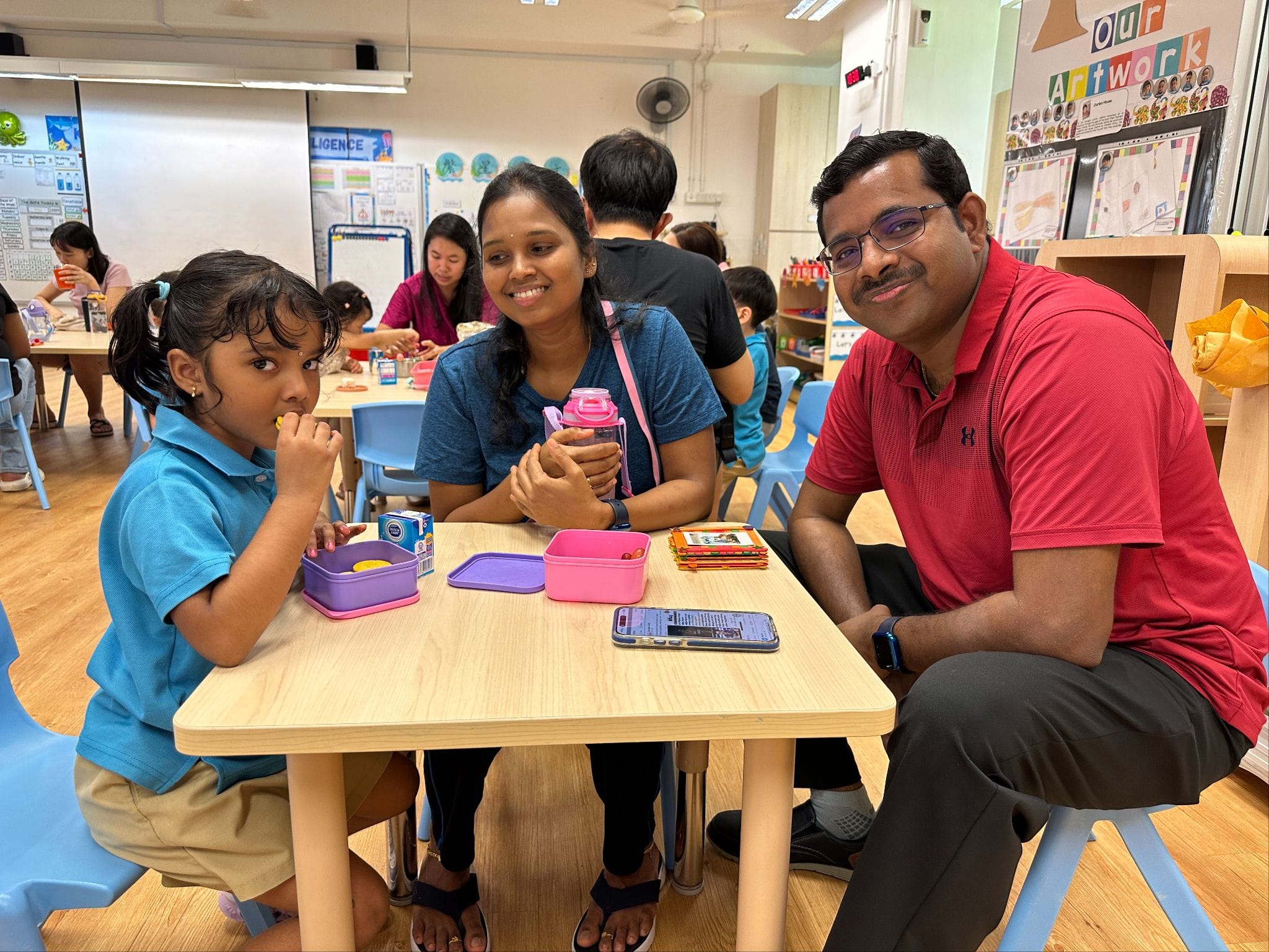 A young girl, a smiling woman in blue, and a man in red sit together at a small wooden table in a classroom.