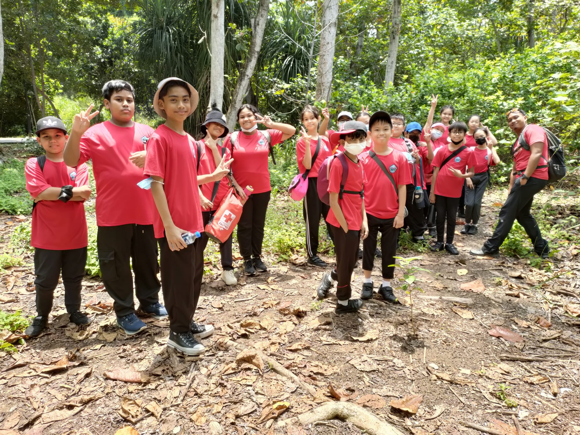 A group of children in matching red shirts pose with peace signs on a forest trail with trees behind them.