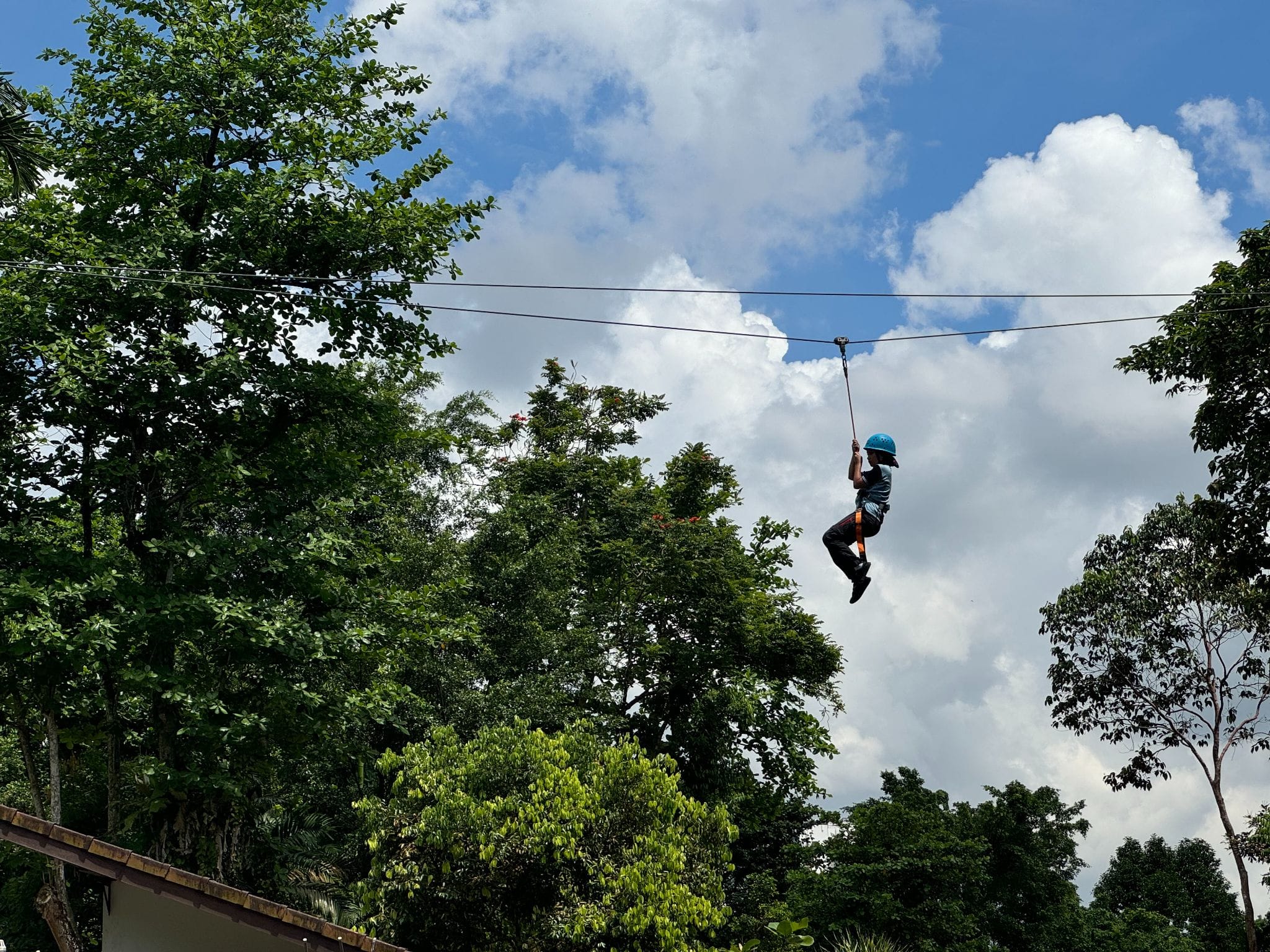 A person wearing a blue helmet rides a zipline high above green trees against a partly cloudy blue sky.