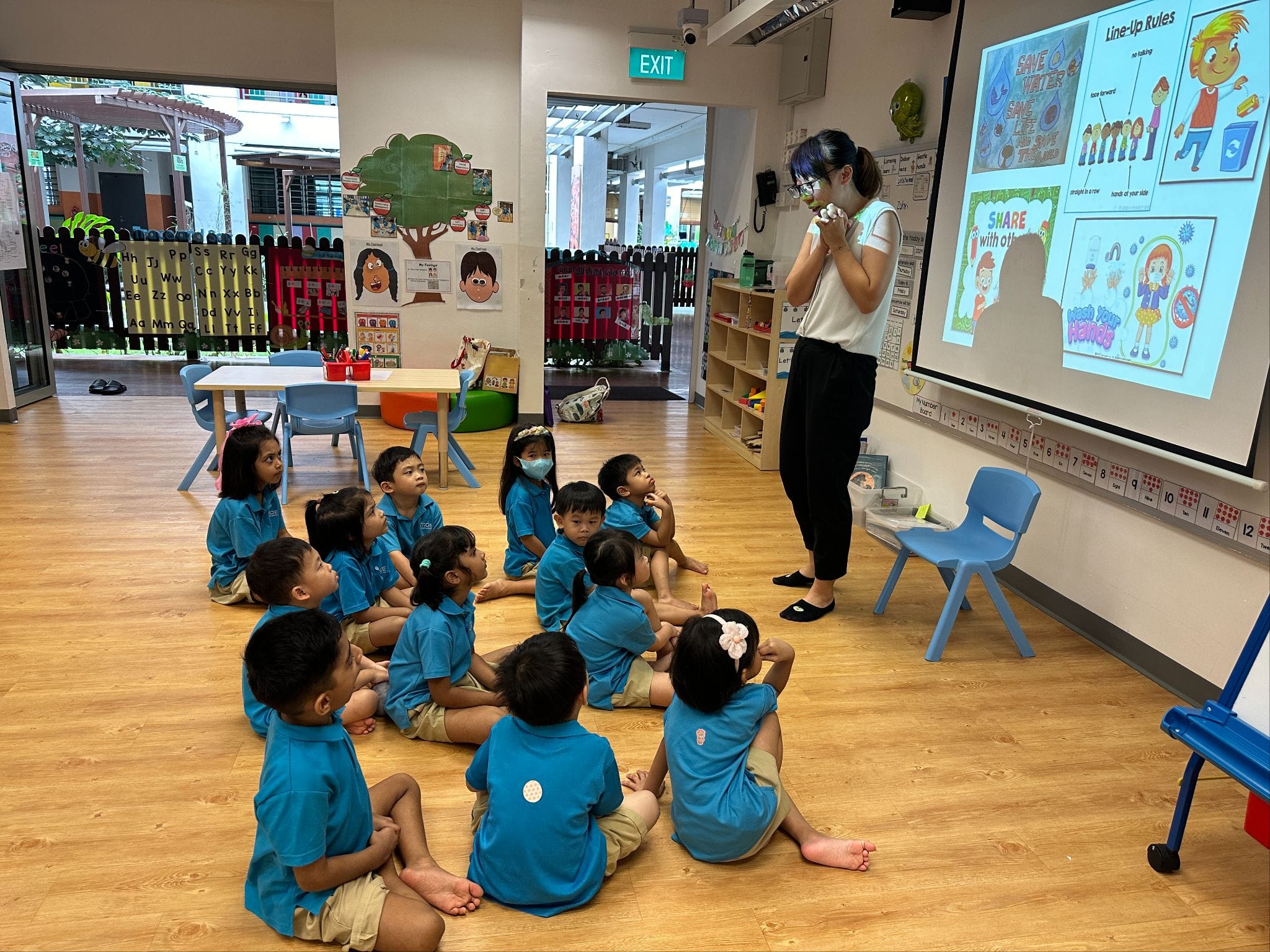 A teacher stands before young children in blue uniforms seated on a classroom floor, with a projected slideshow behind her.
