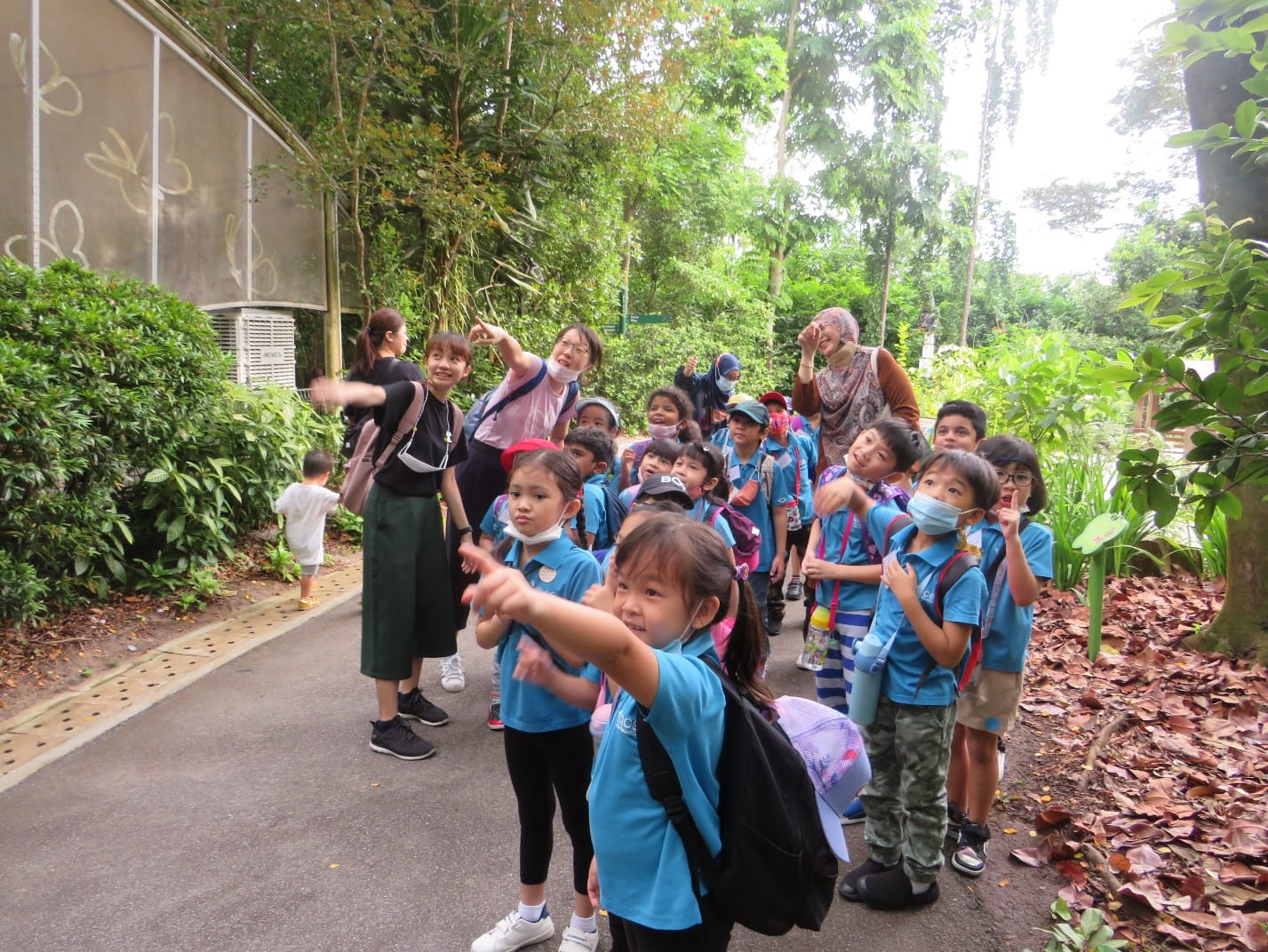 A group of young children in blue uniforms with backpacks on an outdoor field trip, pointing and looking around lush green trees.