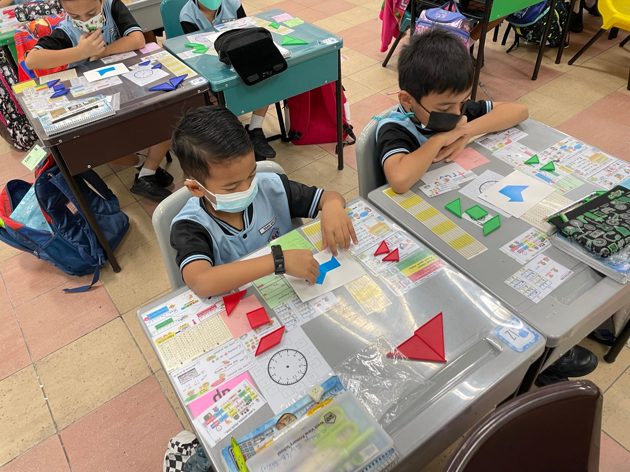 Young students wearing face masks sit at desks arranging colorful paper geometric shapes during a classroom activity.