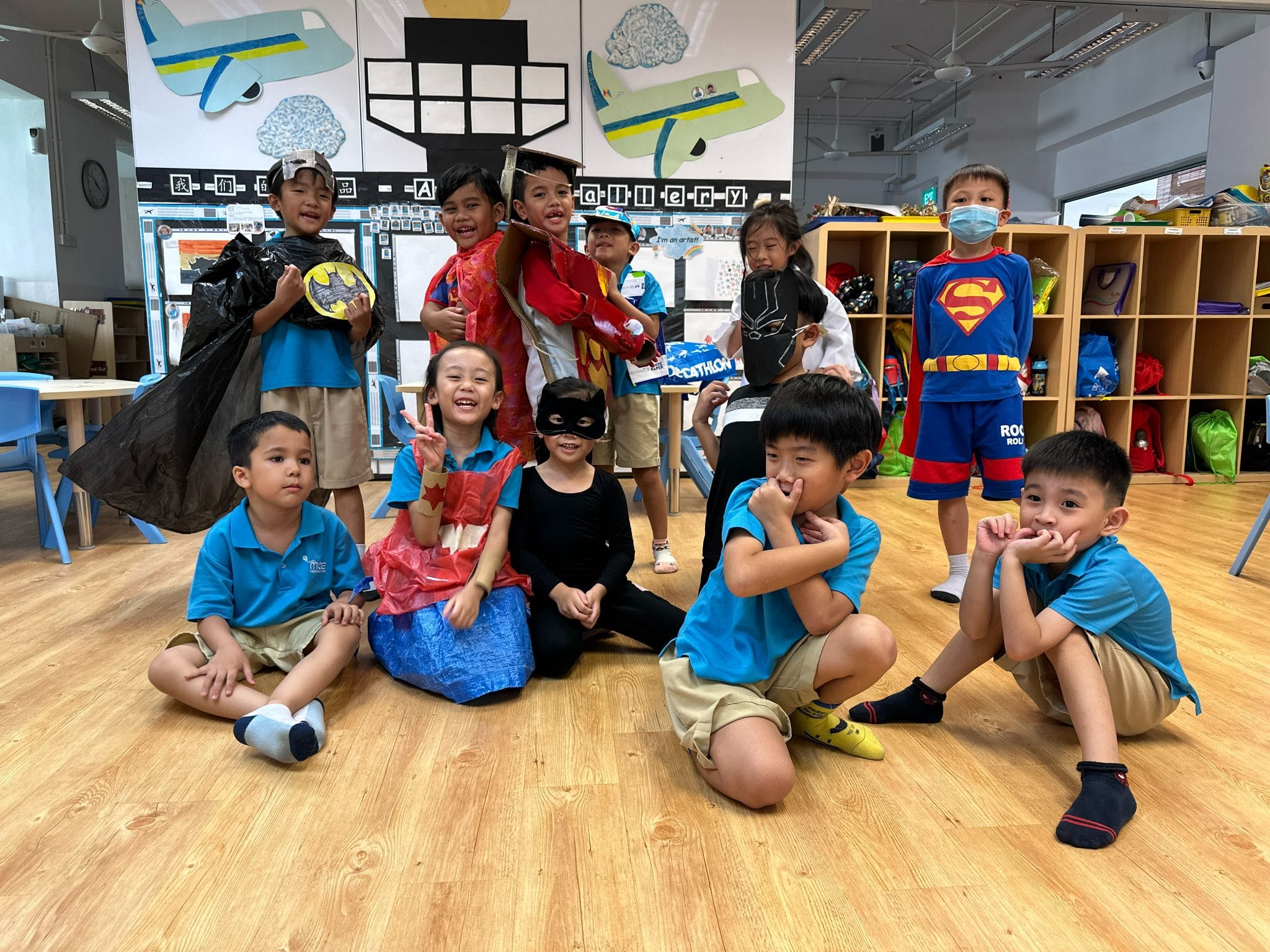 A group of young children in superhero costumes pose and smile in a colorful classroom with airplane decorations.
