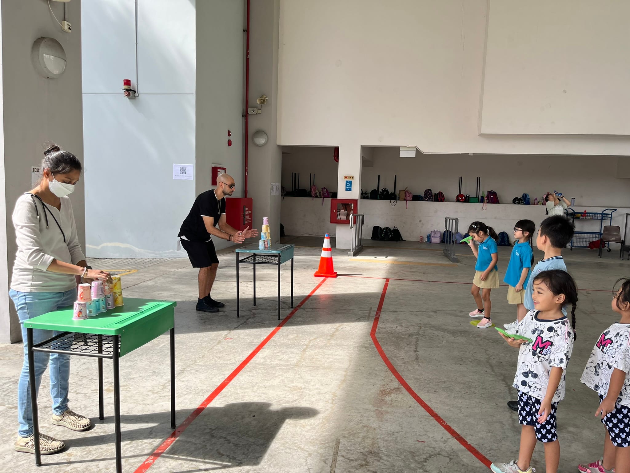 Two adults lead a cup-stacking game activity for young children in a large indoor hall with red line markings.